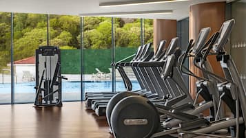 Une rangée de vélos d’appartement et un appareil d’aviron dans une salle de sport donnant sur une piscine et sur un espace verdoyant à l’extérieur.