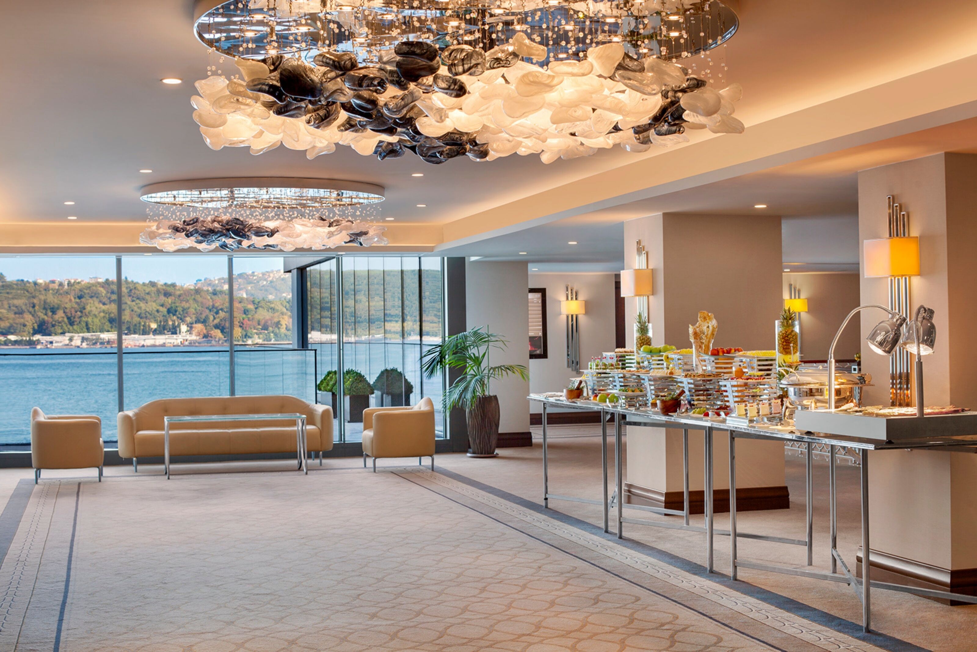 A spacious room with a view of the sea, a buffet table with various food items, and a unique ceiling chandelier.
