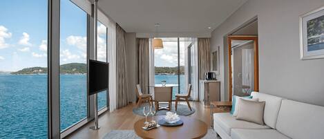 A modern hotel room with a large window overlooking the sea, a dining area with a table and chairs, and a living area with a sofa and a coffee table.