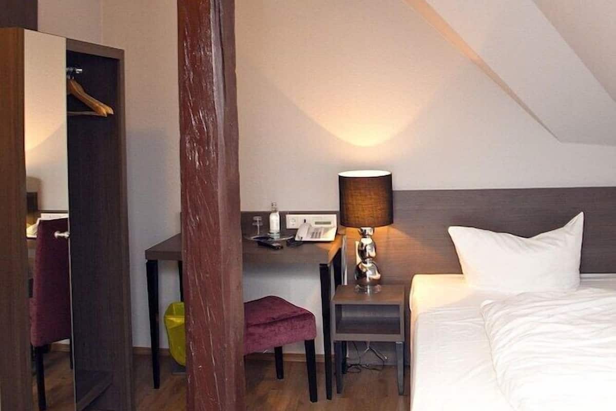 Ein modernes Hotelzimmer mit Holzbalkendecke, einem Bett, einem Schreibtisch mit Lampe, einem Sessel und einem Spiegel.