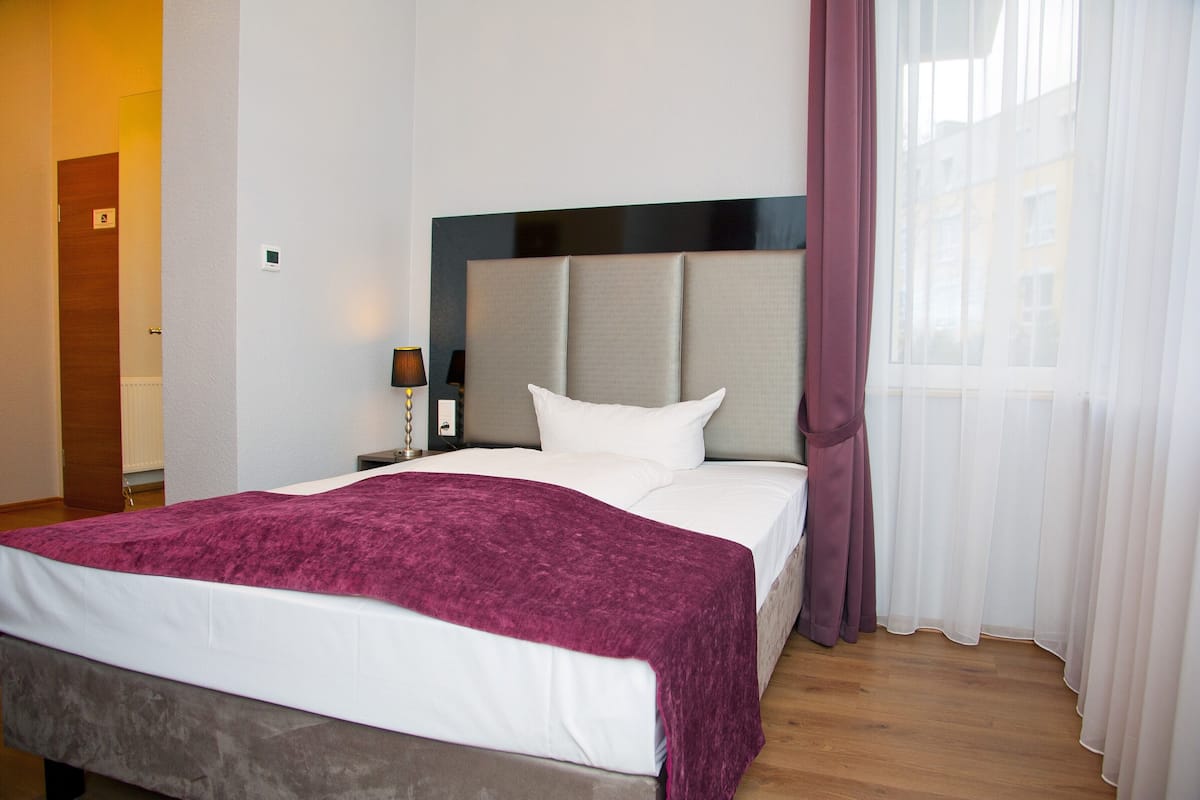 Ein Hotelzimmer mit Bett, Nachttisch, Lampe und einem Fenster mit Vorhängen.