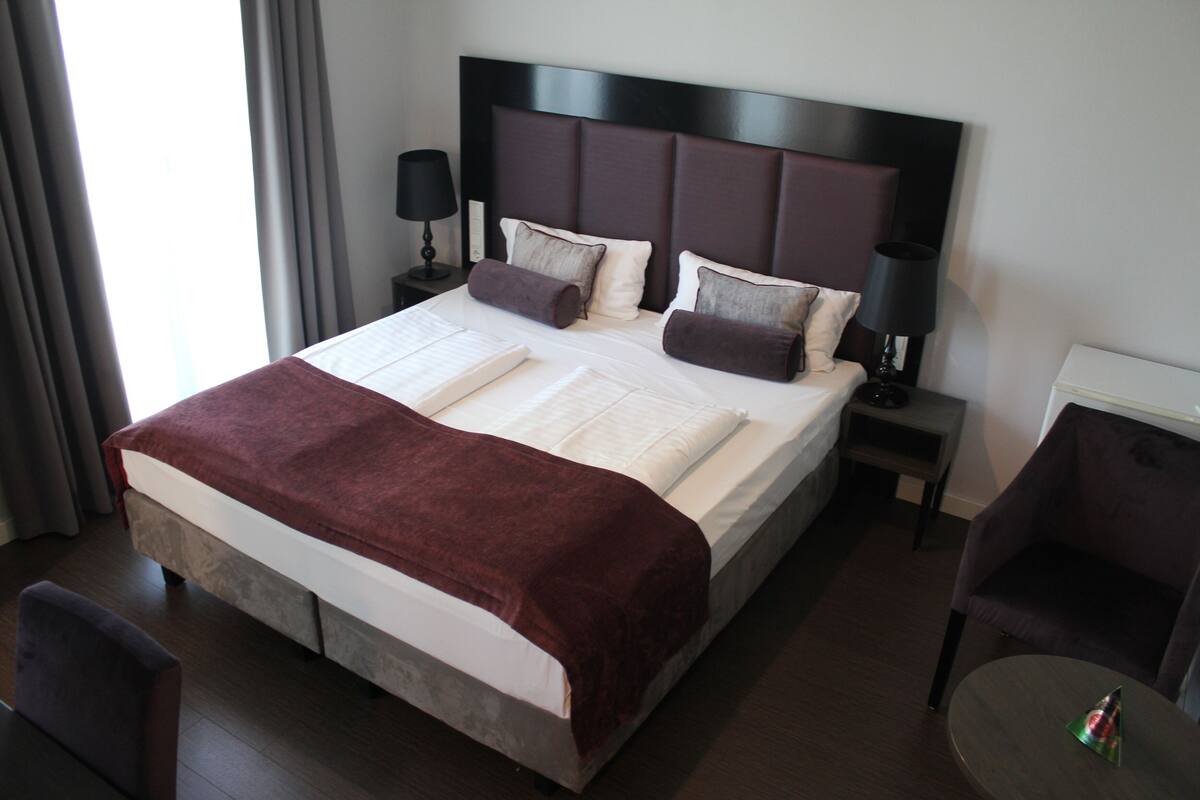 Ein Hotelzimmer mit einem großen Bett, einem Nachttisch mit Lampe, einem Sessel und einem kleinen Tisch mit einer Partyhaube.