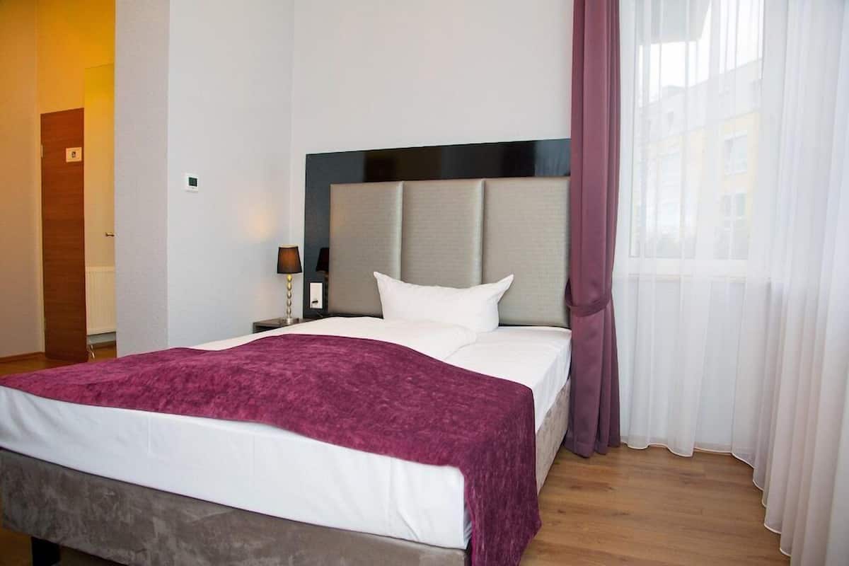 Ein Hotelzimmer mit einem Bett, einem Nachttisch, einer Lampe und einem Fenster mit Vorhängen.