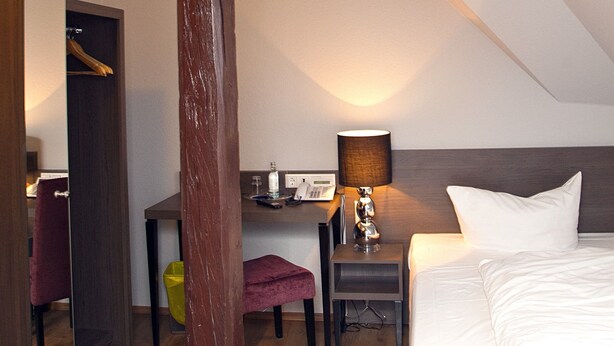 Een slaapkamer met een houten balk in het plafond, een nachtkastje met een lamp, een bed en een klein bureau met een stoel.