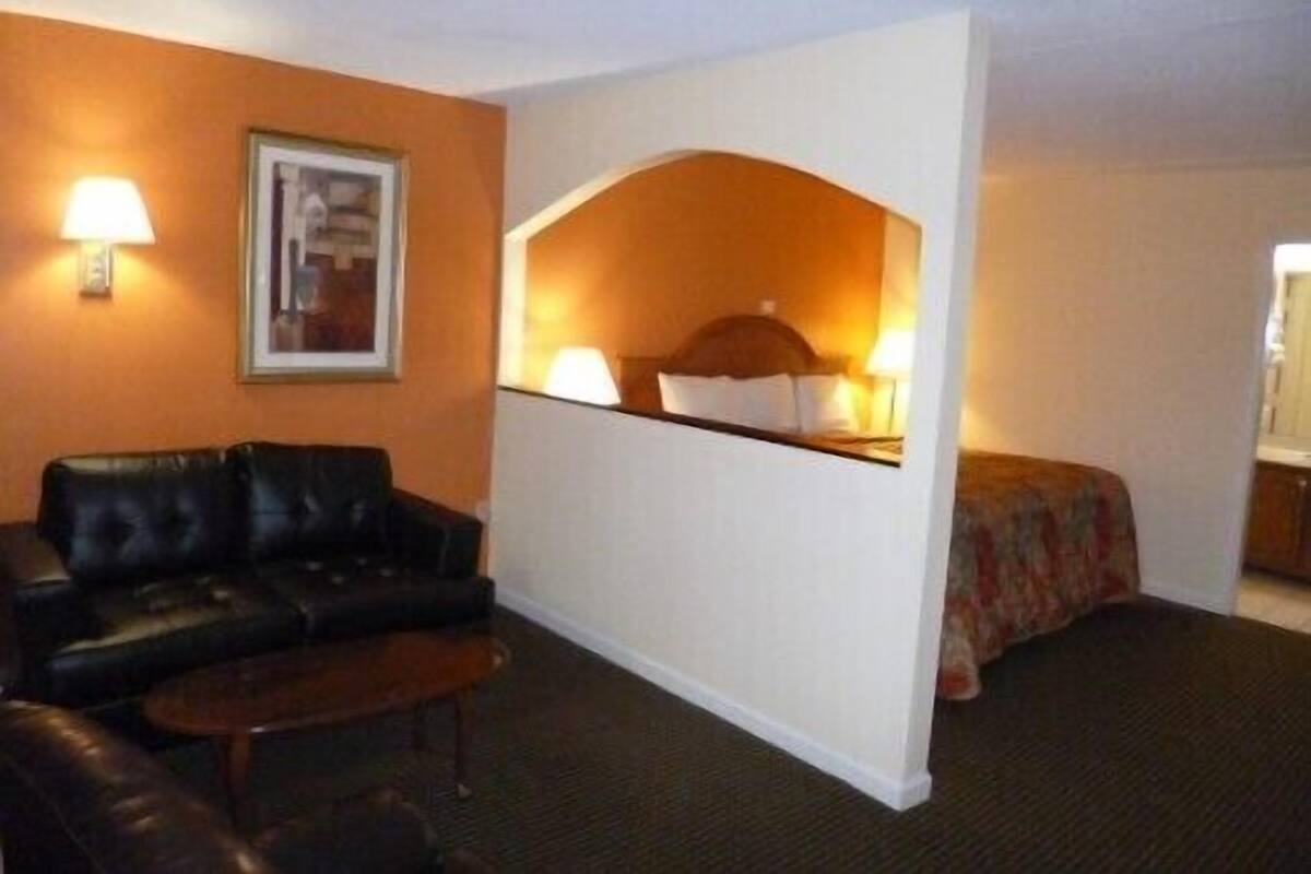 A hotel room with a black leather sofa, a small wooden table, a white partition, and a bed with a floral bedspread.