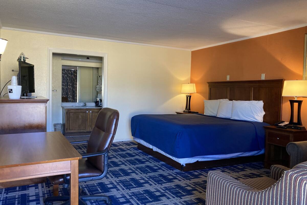 A hotel room with a bed, a desk, a chair, a small table, a lamp, and a nightstand.