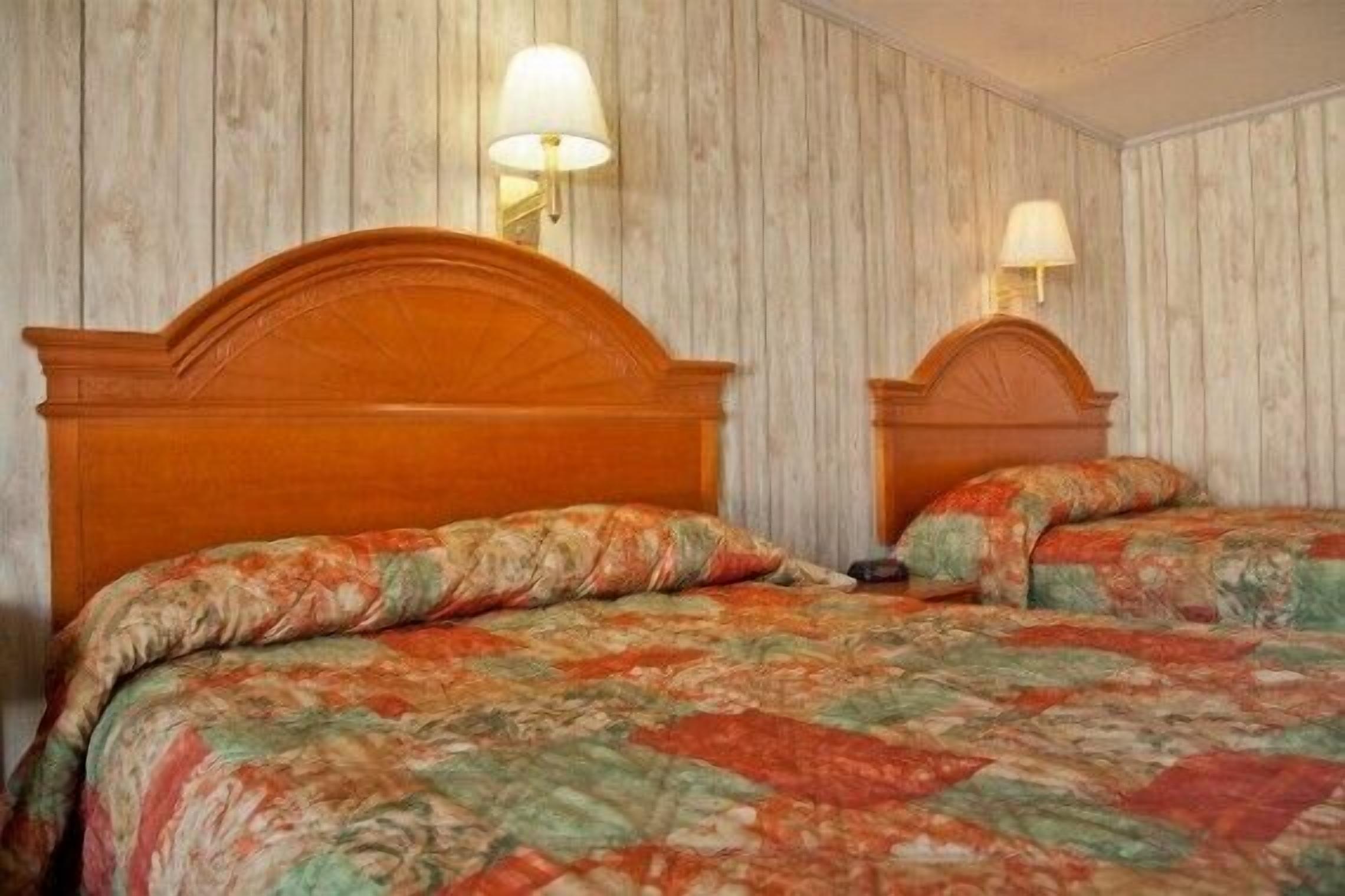 Cama doble con edredón estampado de flores en una habitación con paneles de madera y dos lámparas fijadas a la pared.