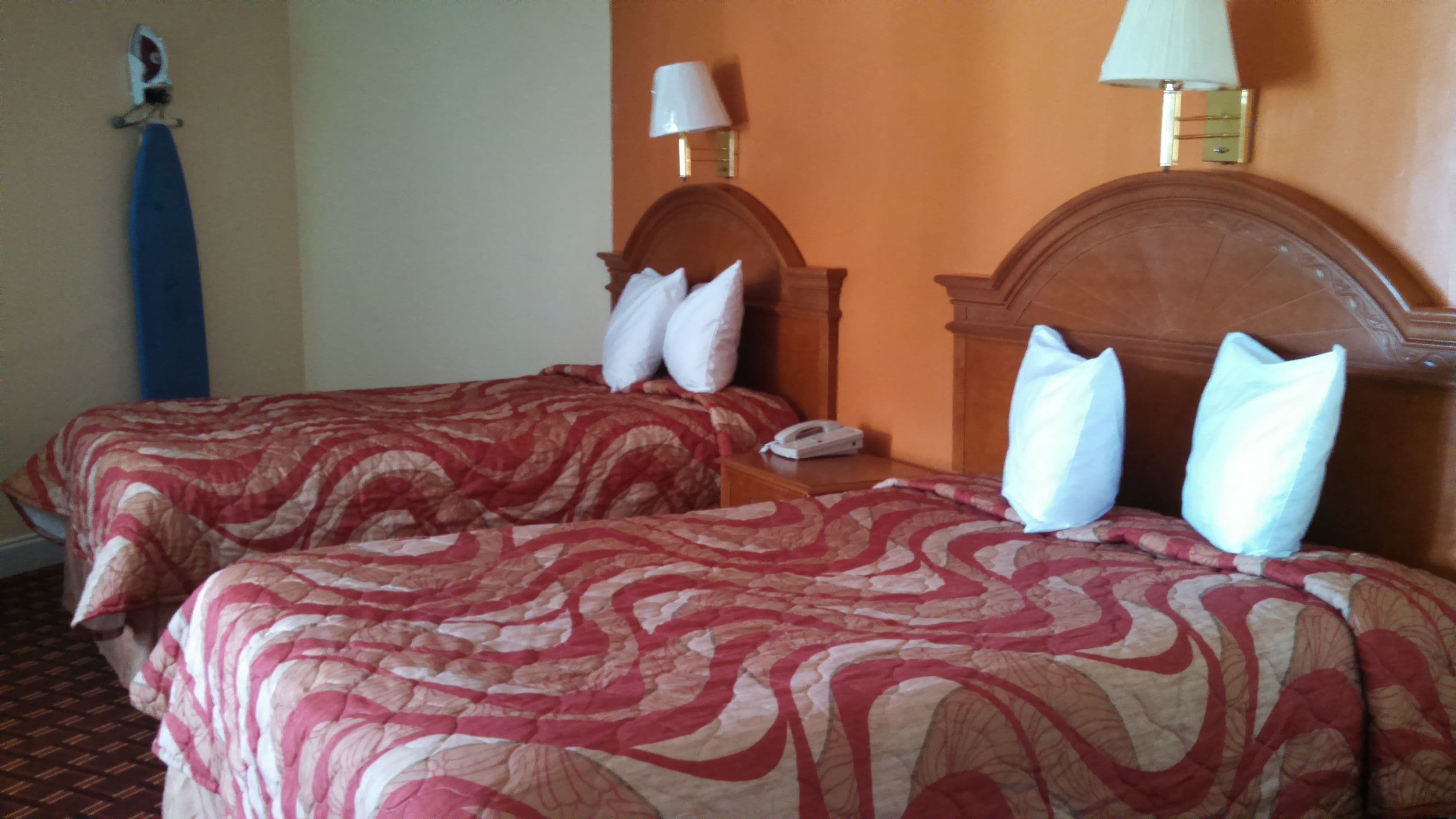 Ein Hotelzimmer mit zwei Betten, jeweils mit einem hölzernen Kopfteil und roten, gemusterten Bettdecken. Es gibt zwei Nachttische mit Lampen und einem Telefon.