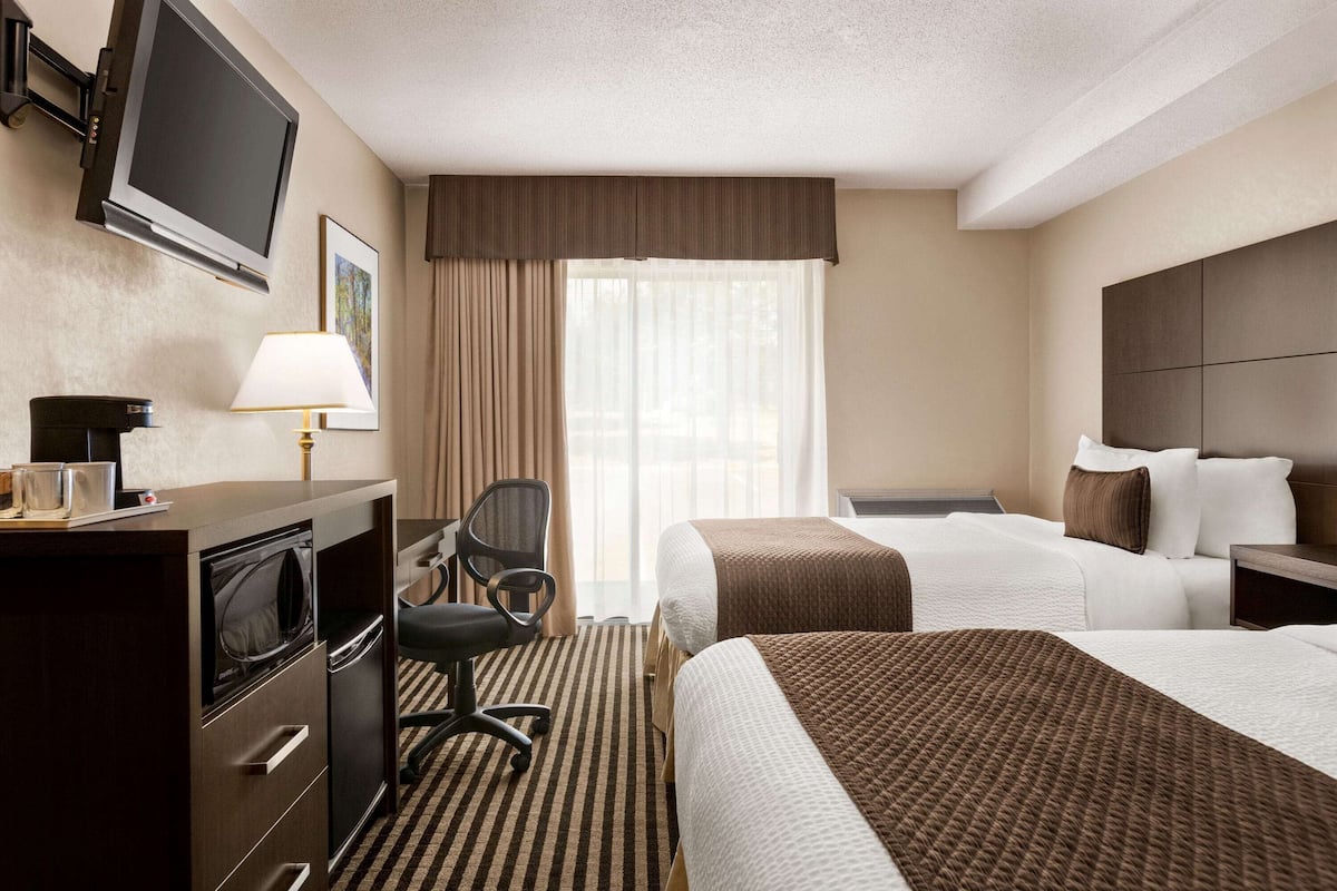 A hotel room with a flat-screen TV, a desk with a lamp, a chair, two beds with brown and white bedding, and a large window with curtains.