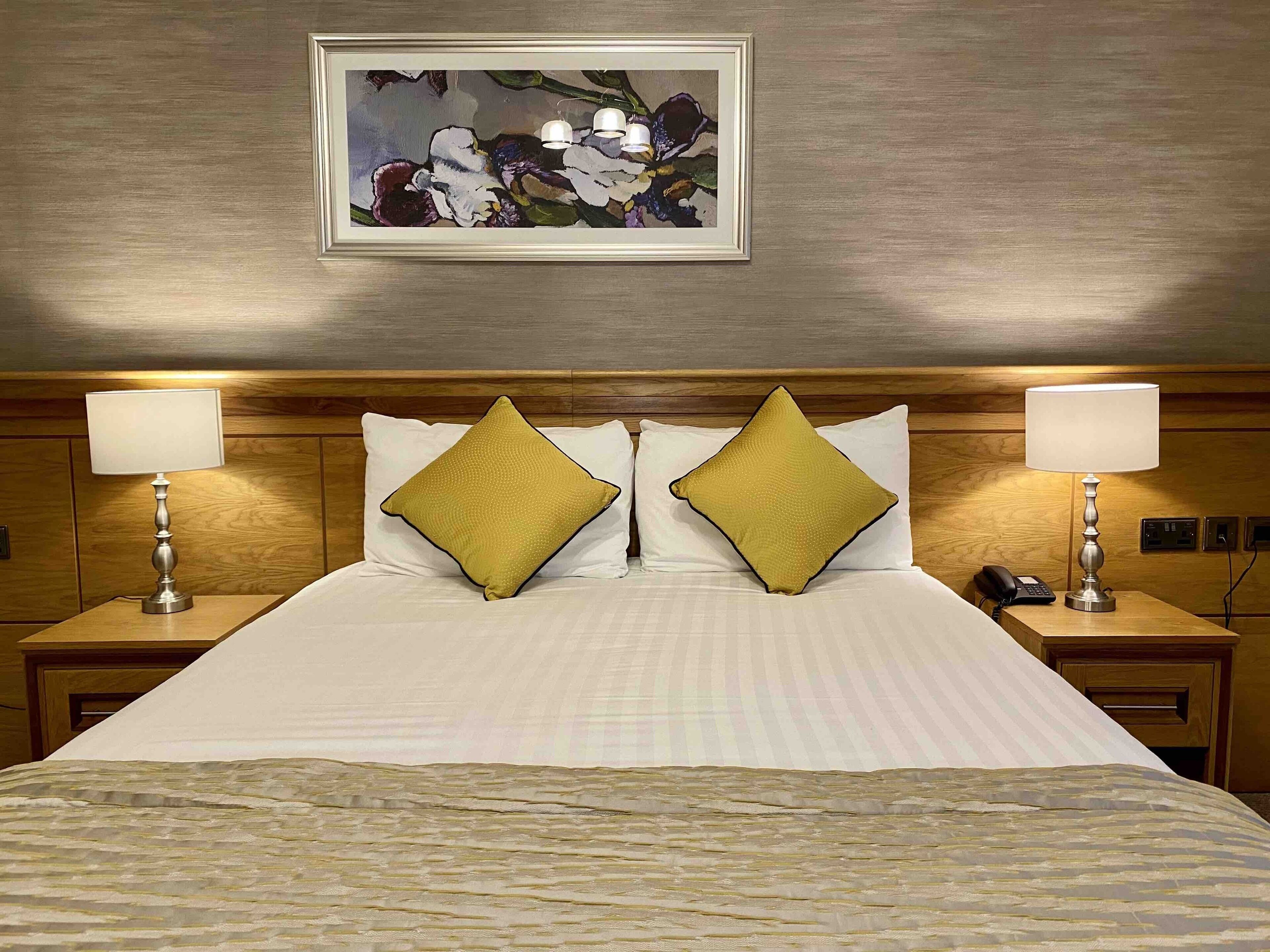 Habitación de hotel con una cama grande, dos mesitas de noche, dos lámparas y una obra enmarcada sobre la cama.