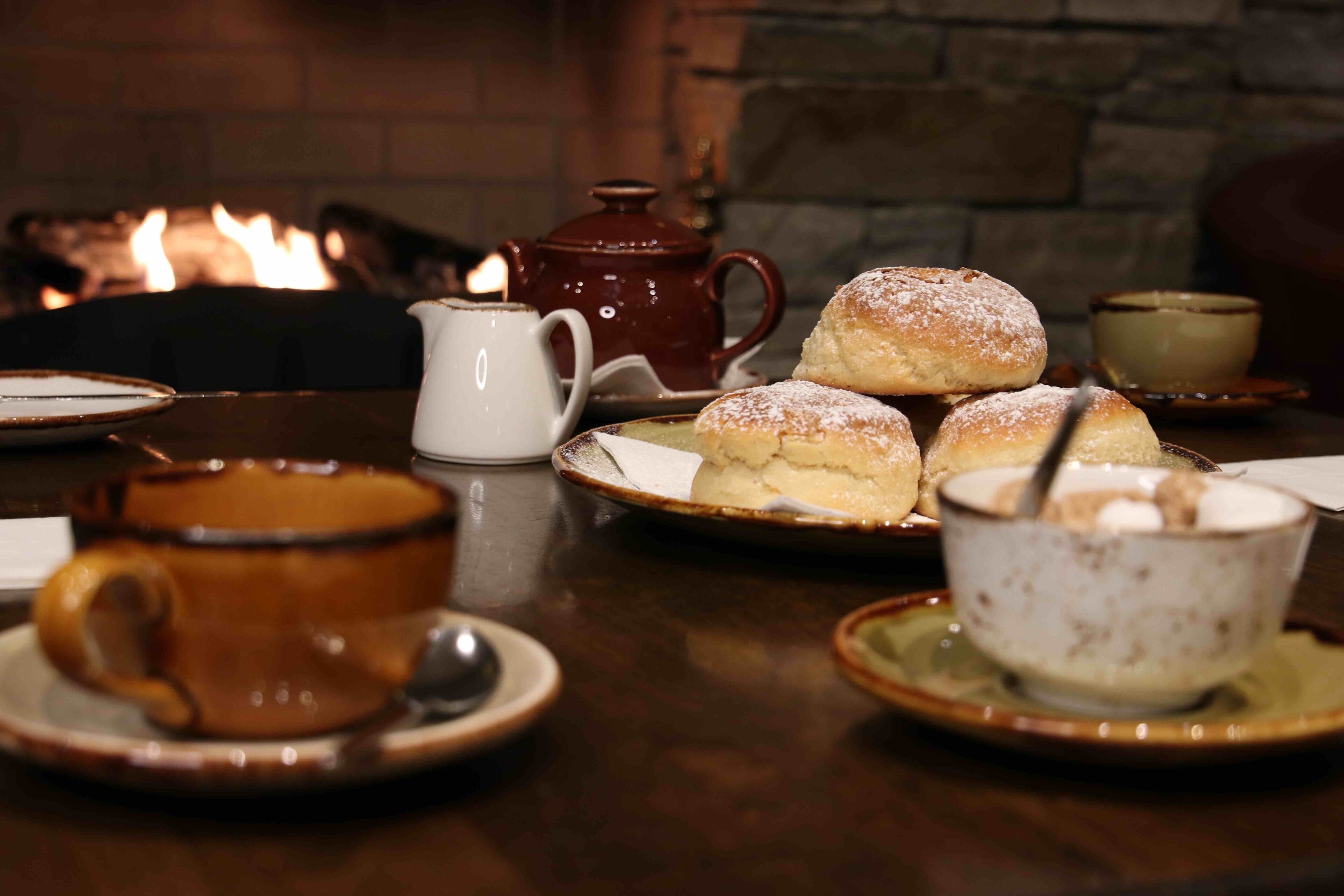 Una mesa servida con pasteles, una tetera y tazas, con una chimenea al fondo.