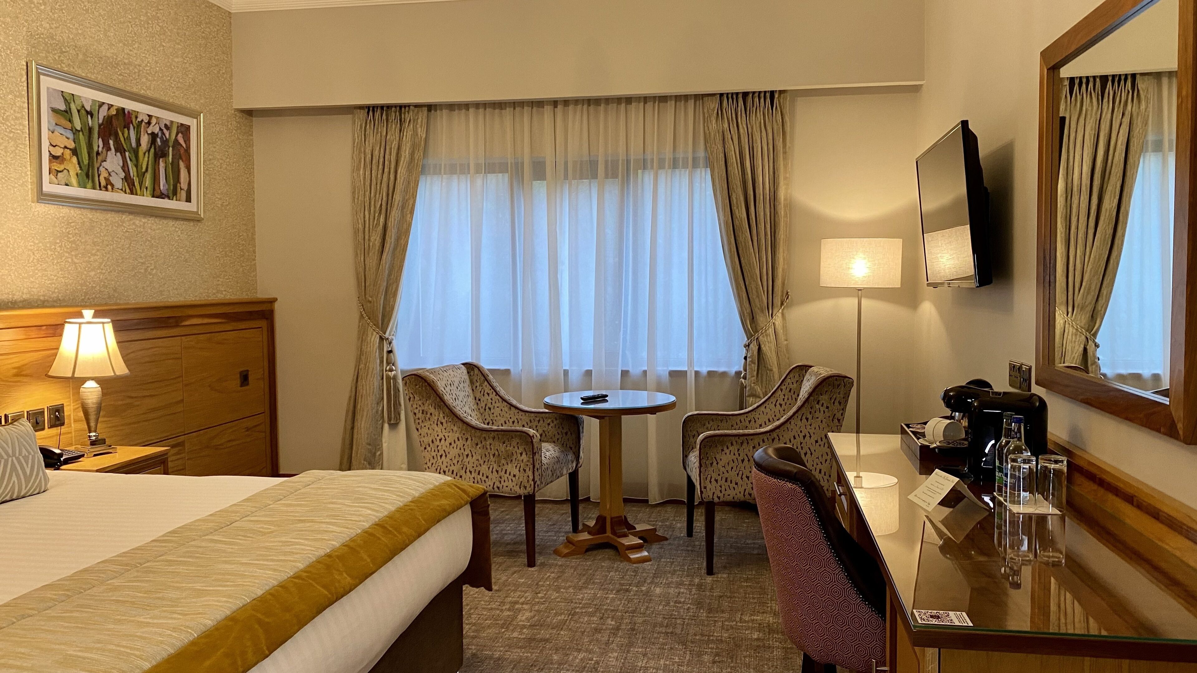 A hotel room with a bed, a desk with a chair, a small table, a TV, and a window with curtains.