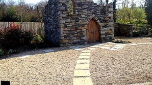 A stone structure with a wooden door, surrounded by gravel and a stone pathway. - An Grianan Hotel (Burt)