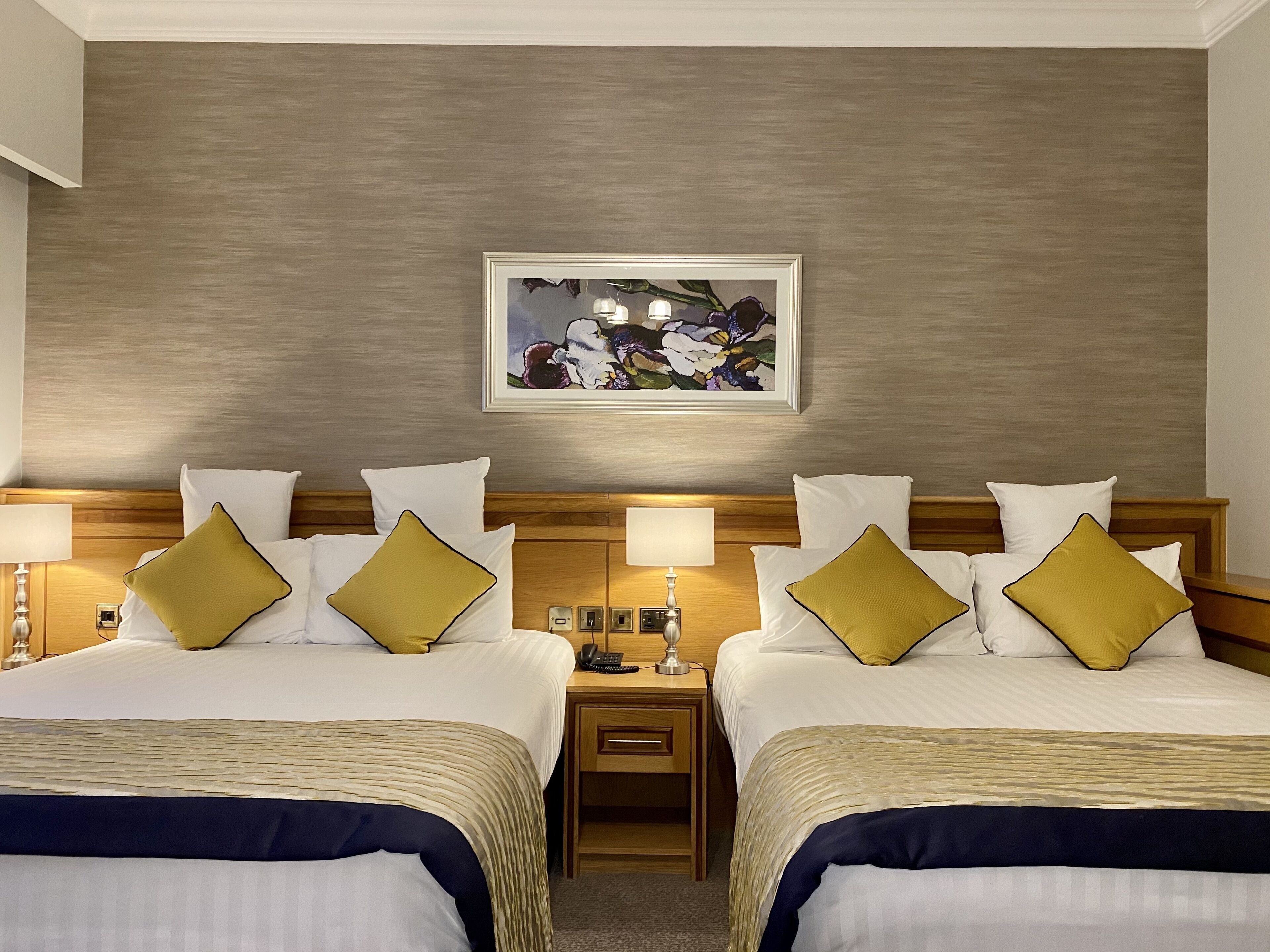 A hotel room with two beds, each with white bedding and gold pillows, a wooden headboard, a nightstand with a lamp, and a framed floral artwork on the wall.
