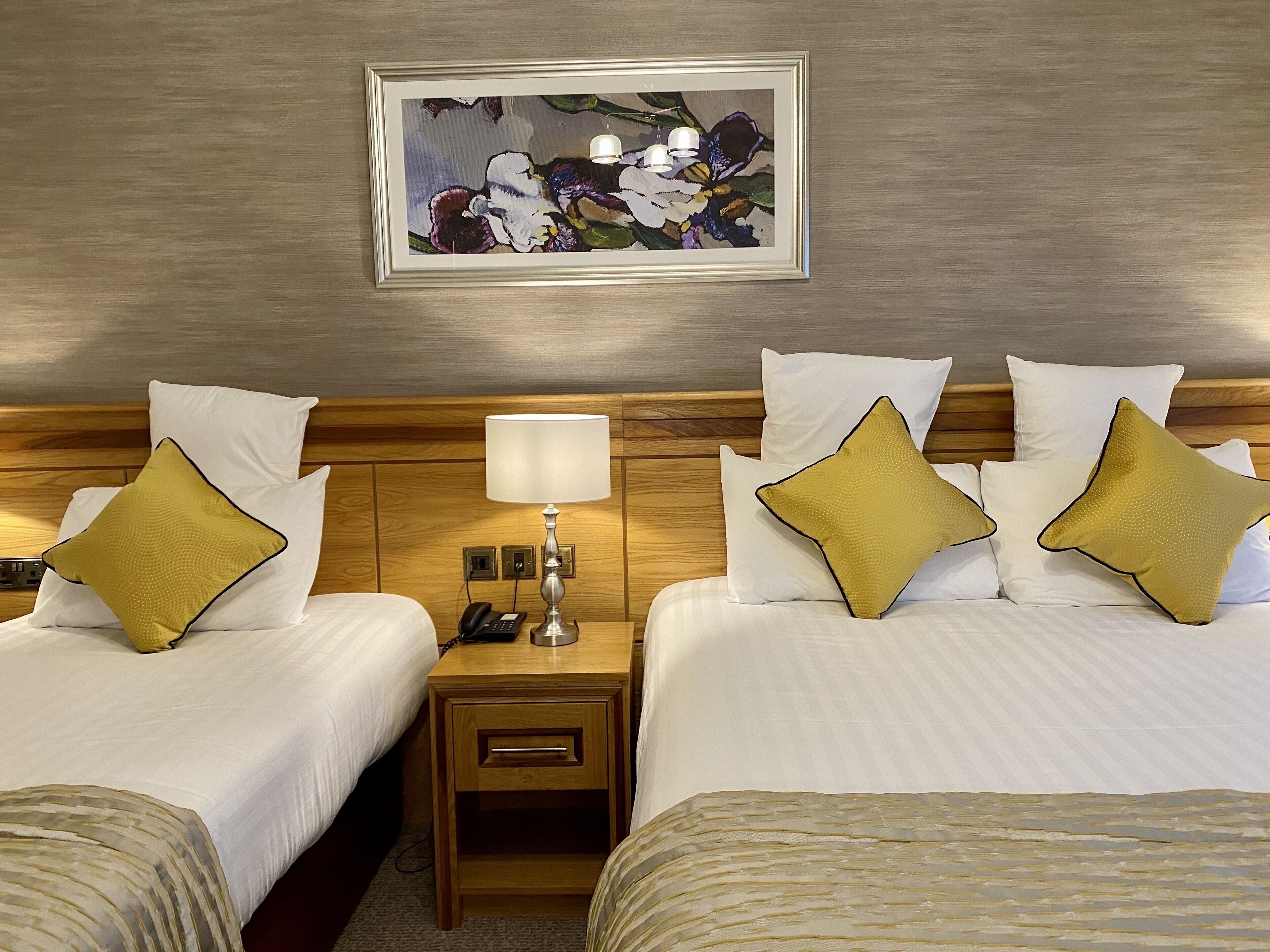 A hotel room with two beds, each with white bedding and gold pillows, a bedside table with a lamp, a framed artwork on the wall, and a wooden headboard.