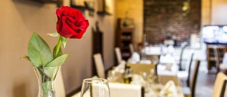 Una mesa de comedor con un florero que contiene una rosa roja, dos copas de vino vacías y un mantel blanco.