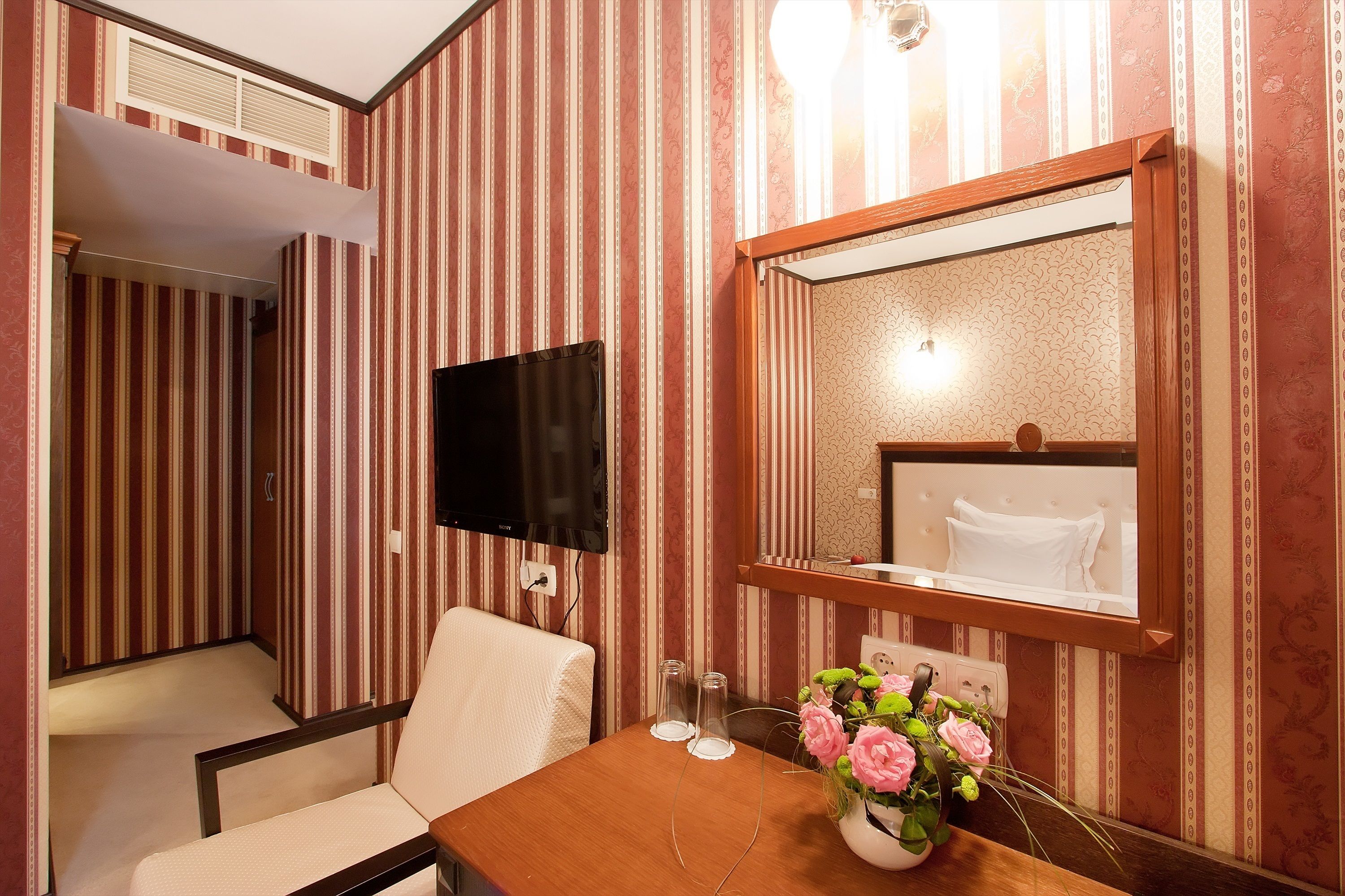 A hotel room with striped wallpaper, a wooden table with a vase of flowers, a chair, a flat-screen TV, and a mirror above a bed.