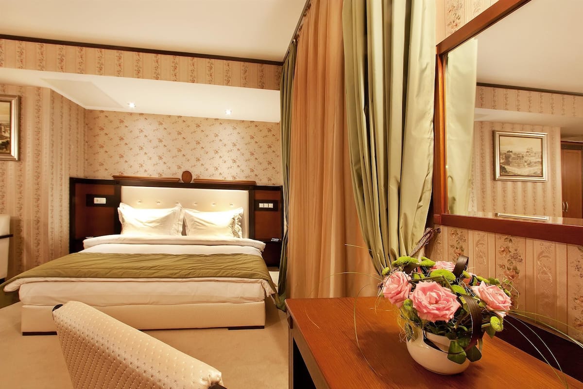 A hotel room with a neatly made bed, a wooden nightstand with a vase of flowers, and a large mirror reflecting the room.