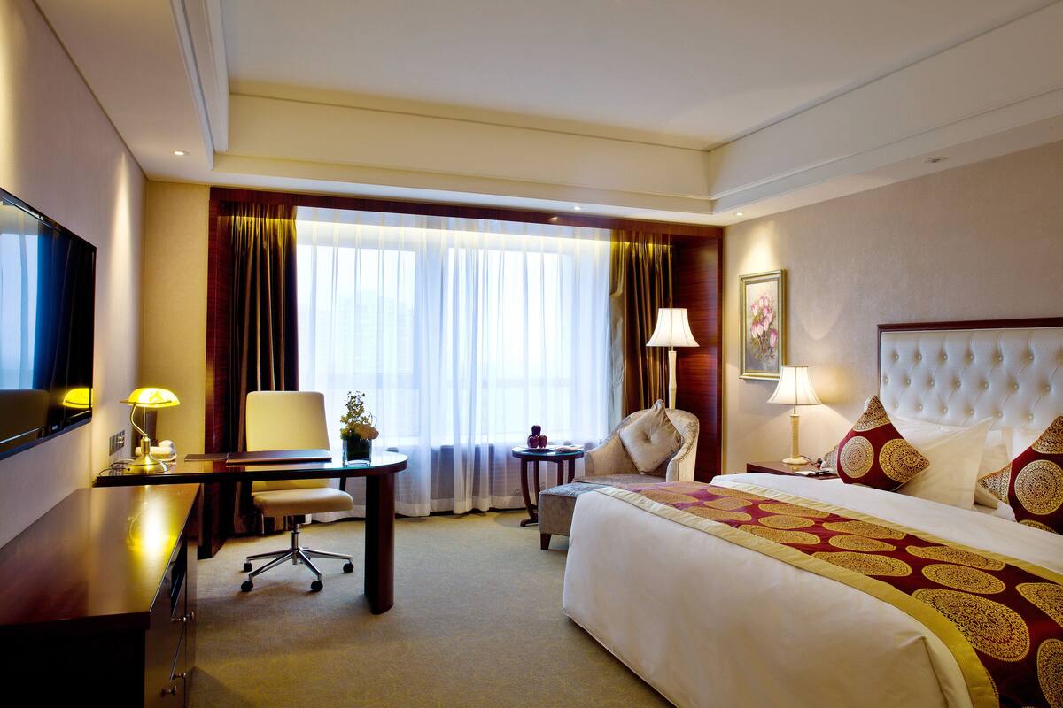 A hotel room with a large bed, a desk, a chair, a TV, and a window with curtains.