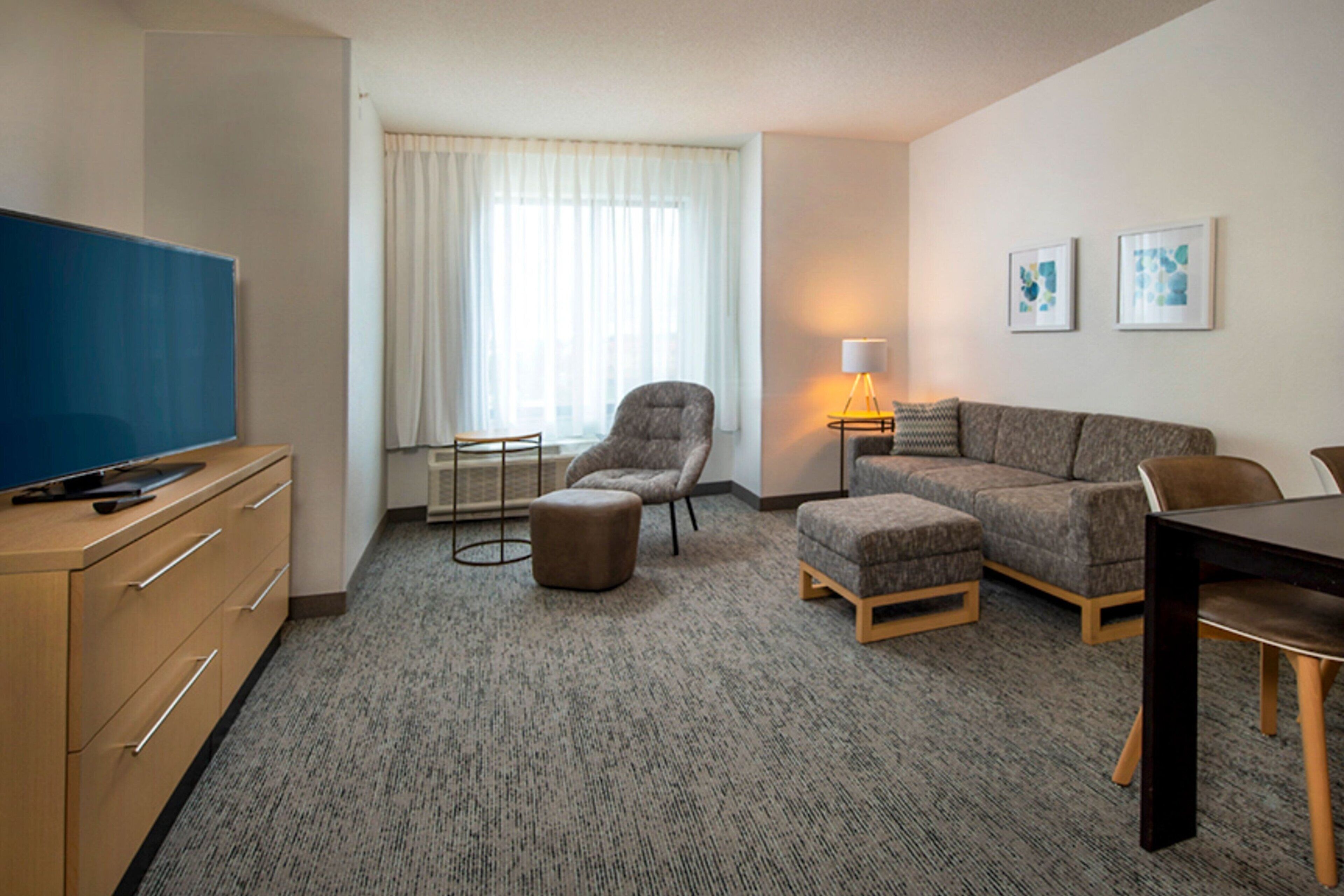 Hotelkamer met flatscreen televisie, een bank, een stoel, een klein voetenbankje, een lamp en een eettafel.