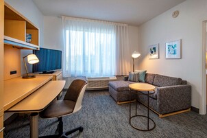 Una habitación de hotel moderna con escritorio, silla, sofá y una pequeña mesa redonda. Hay un televisor sobre un mueble bajo, una ventana con cortinas y dos cuadros en la pared.