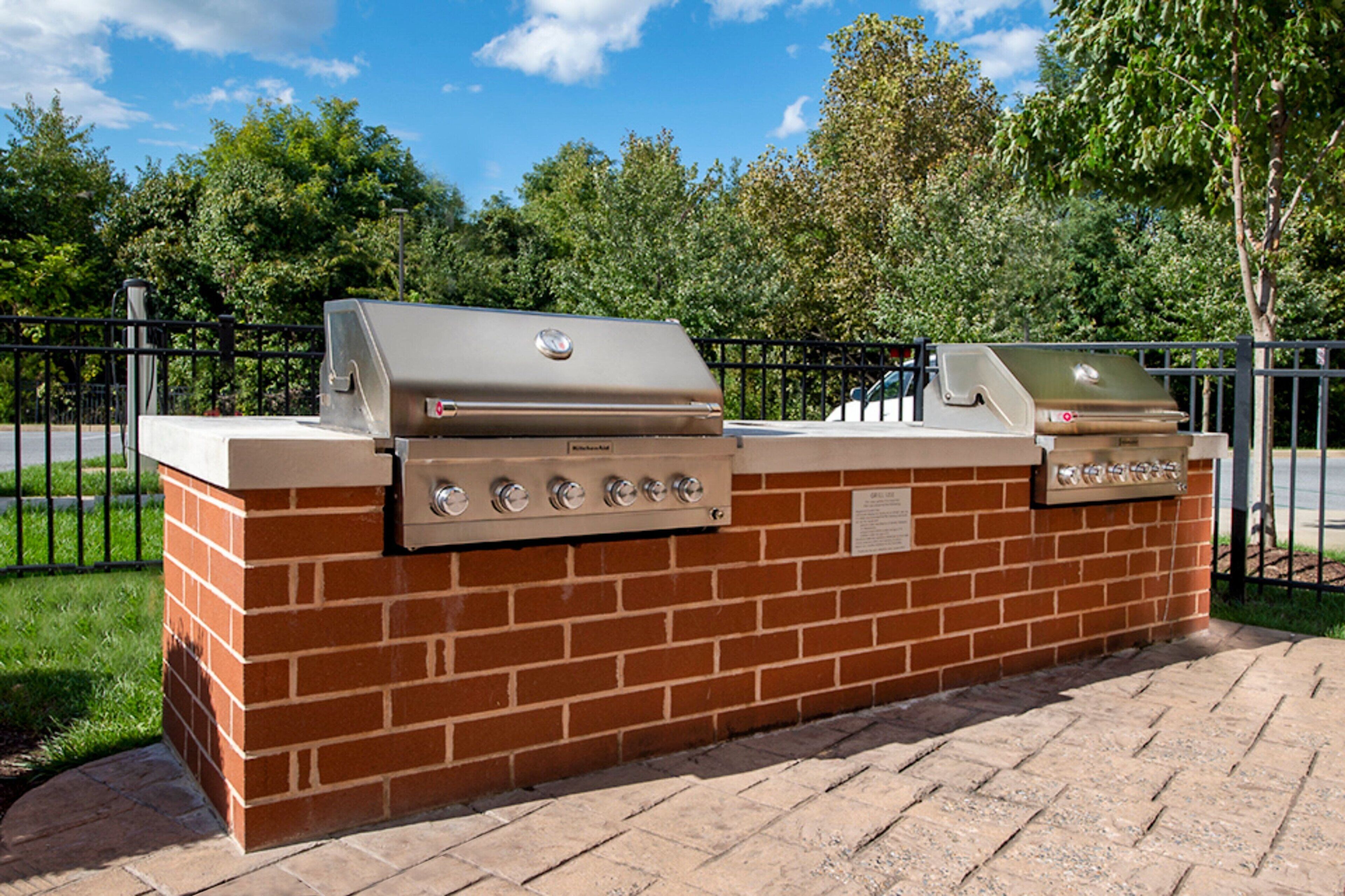 Una parrilla de ladrillo para exteriores con quemador integrado y un quemador lateral, rodeada por una cerca metálica y árboles.