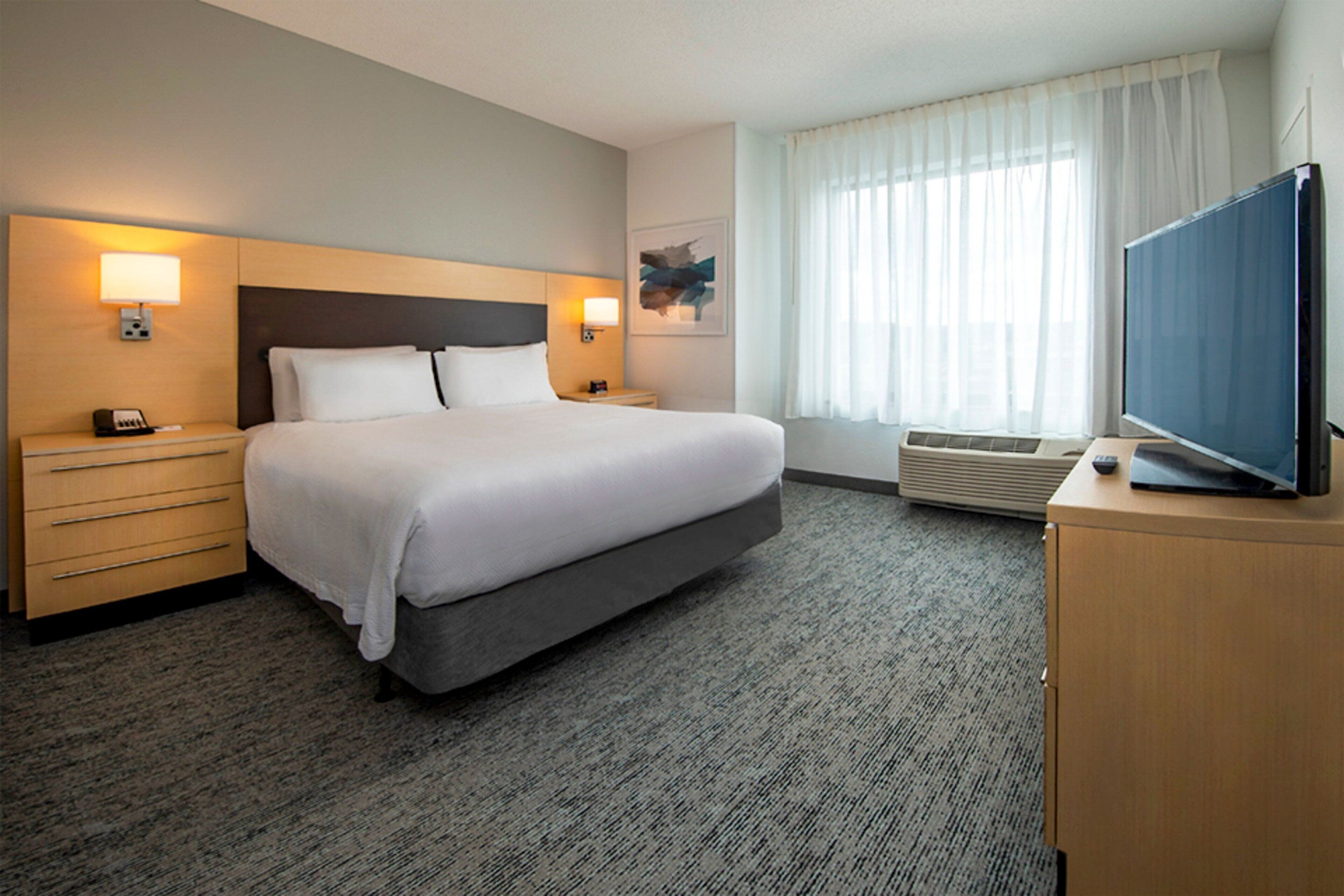 A hotel room with a large bed, two bedside tables with lamps, a flat-screen TV on a wooden dresser, and a large window with sheer curtains.
