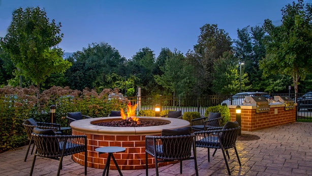 A circular outdoor seating area with a fire pit, surrounded by chairs and tables, set on a brick patio with trees in the background. - TownePlace Suites by Marriott Frederick (Frederick)