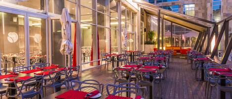 An outdoor dining area with tables and chairs, large windows, and a view of the cityscape.