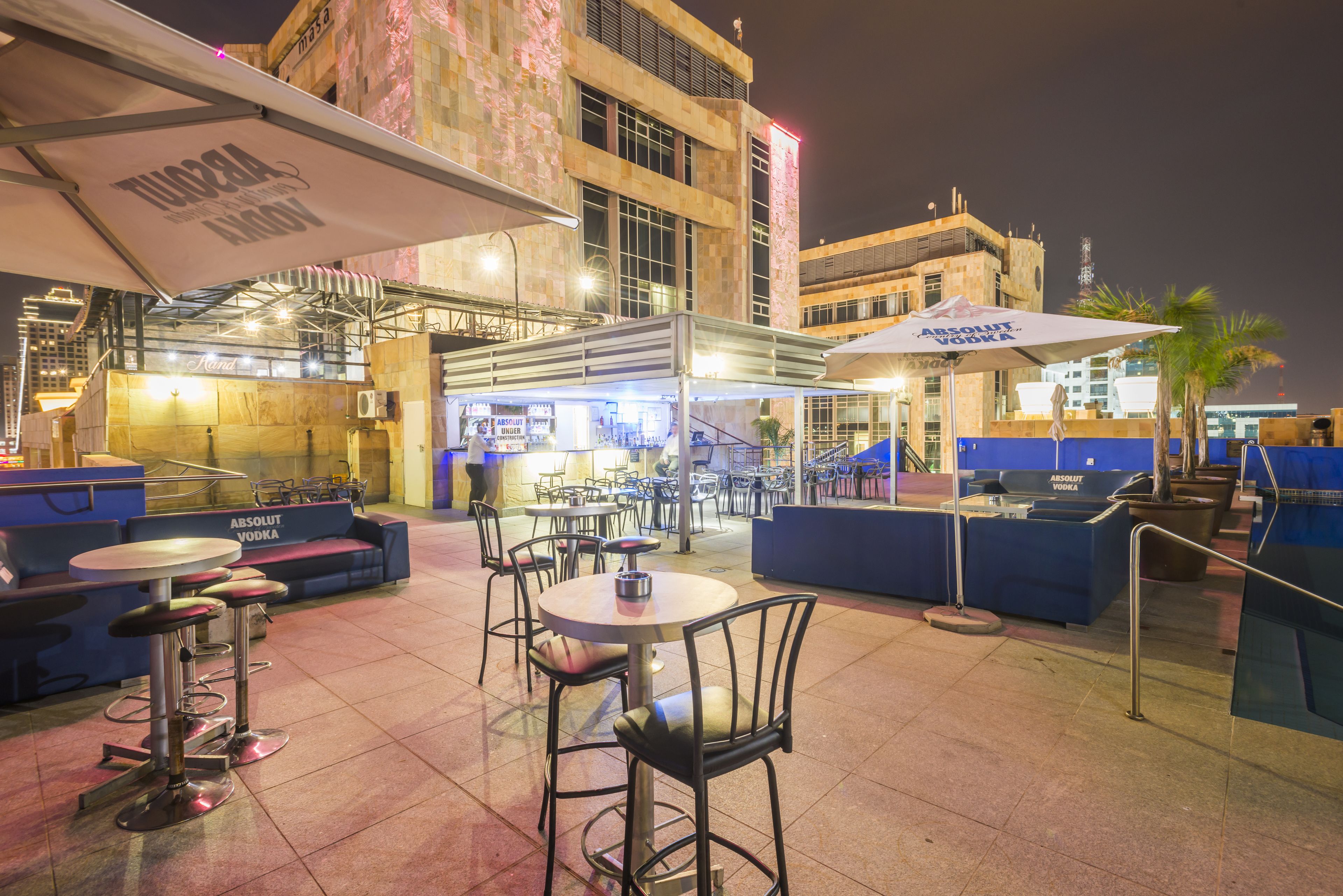 An outdoor dining area with tables and chairs, umbrellas, and a bar area with a sign for Absolut Vodka.