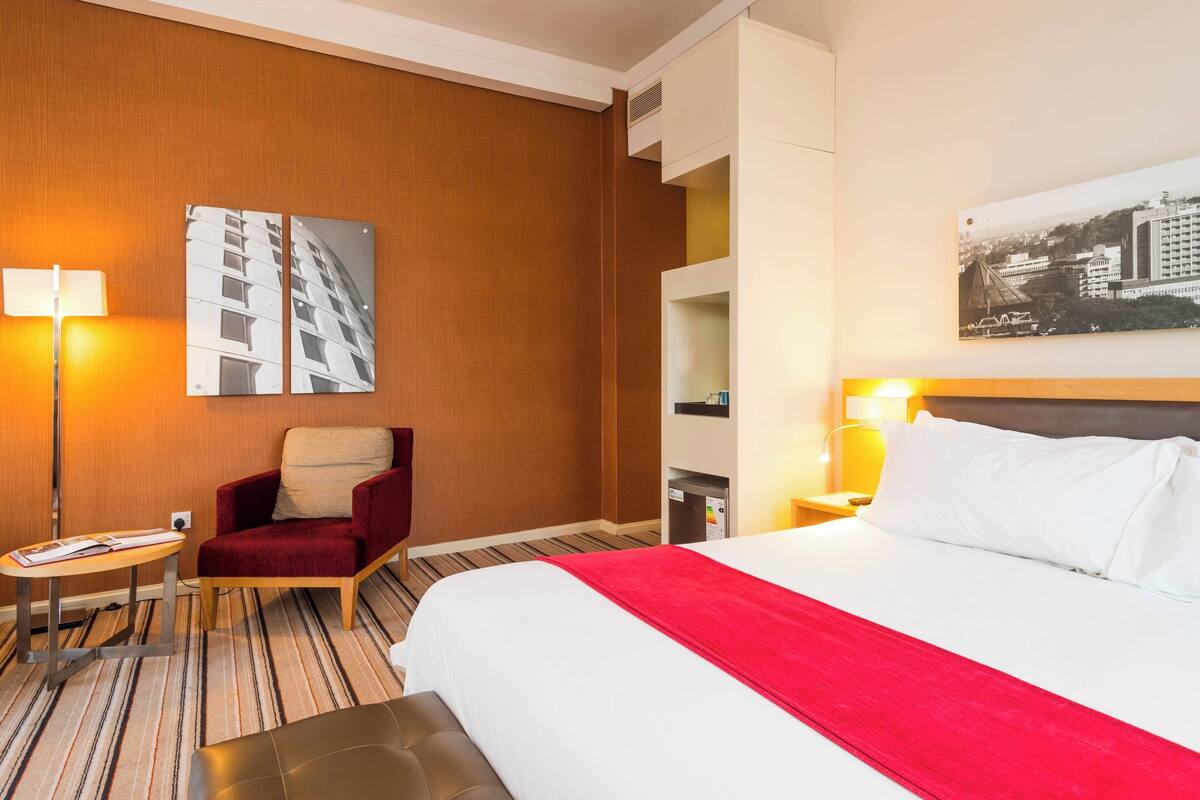 Una habitación de hotel con una cama grande, un sillón rojo, una mesita, un escritorio, una lámpara, una mesita de noche y un estante en la pared.