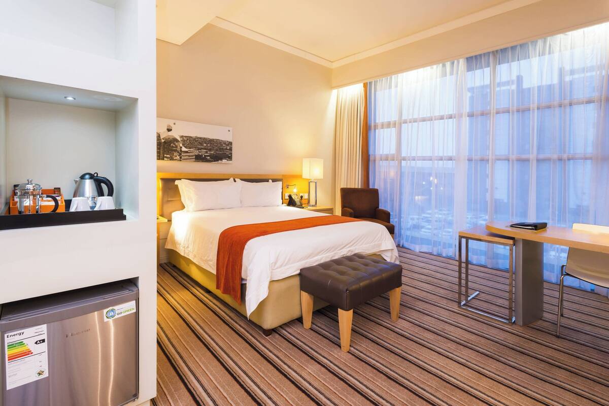 Una habitación de hotel con cama, escritorio, silla, mesita, refrigerador y ventana con cortinas.