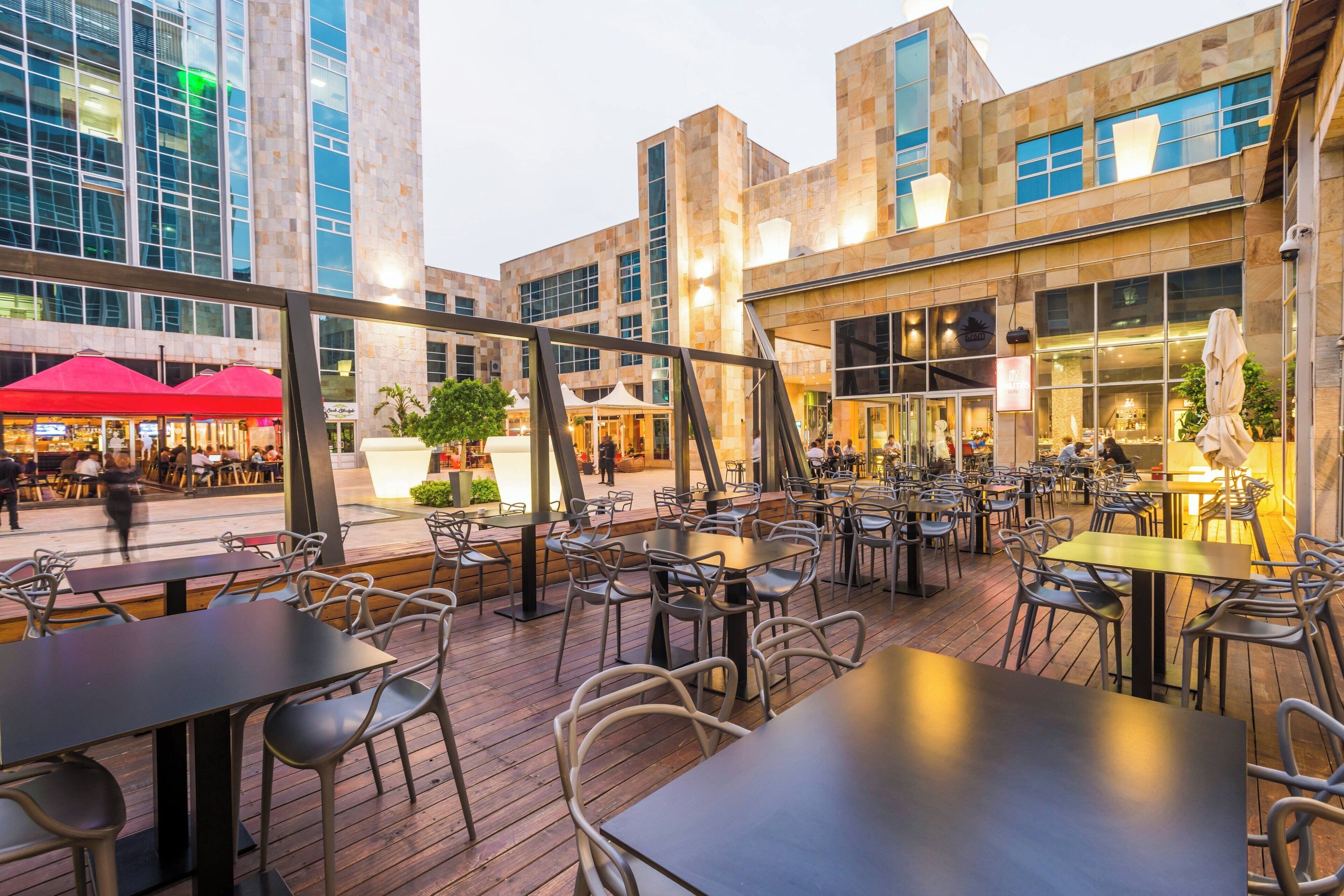 An outdoor dining area with tables and chairs, a wooden deck, and a view of a modern building with large windows and a restaurant in the background.