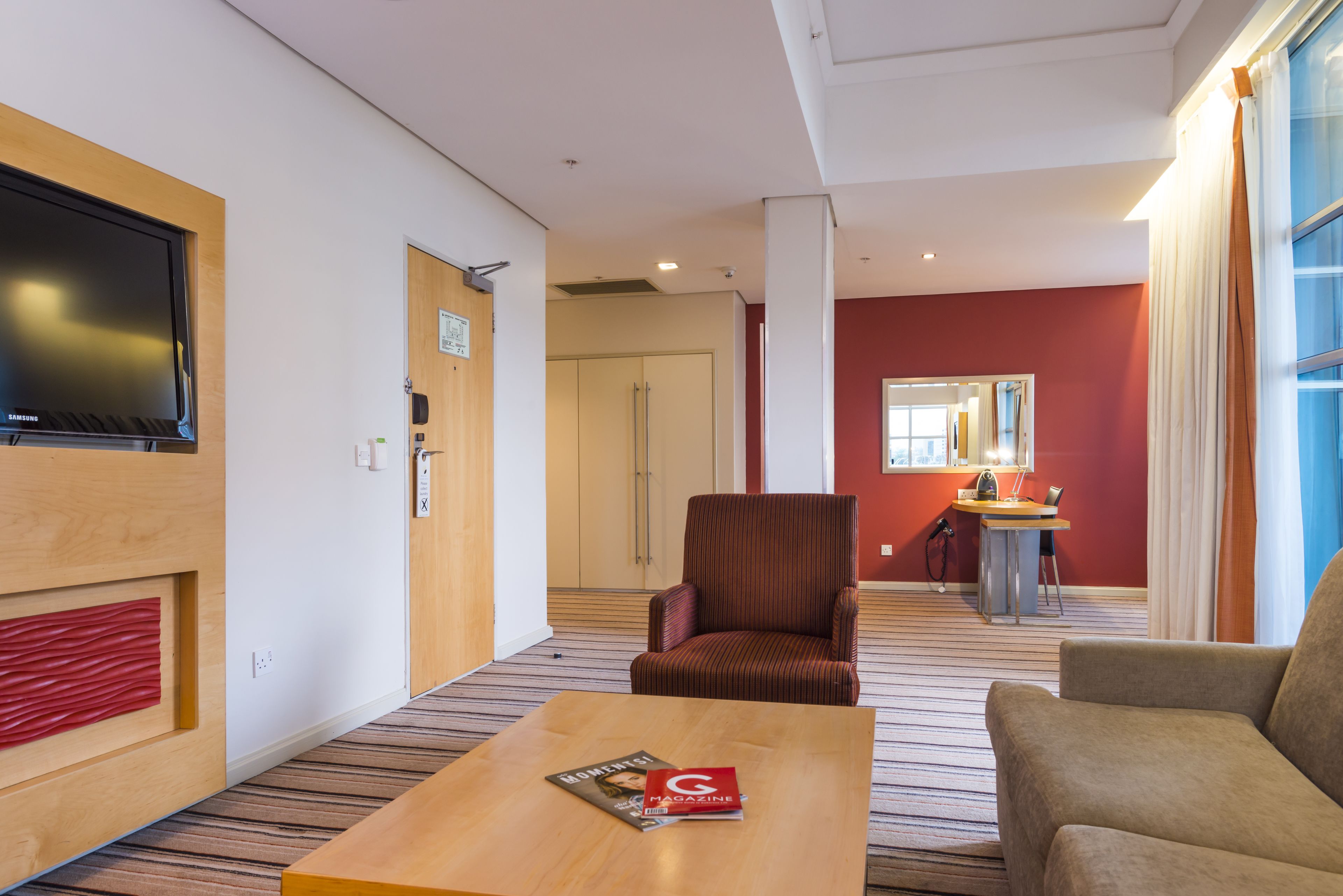 A hotel room with a wooden coffee table, a red armchair, a striped carpet, a television, a wardrobe, and a desk with a lamp.