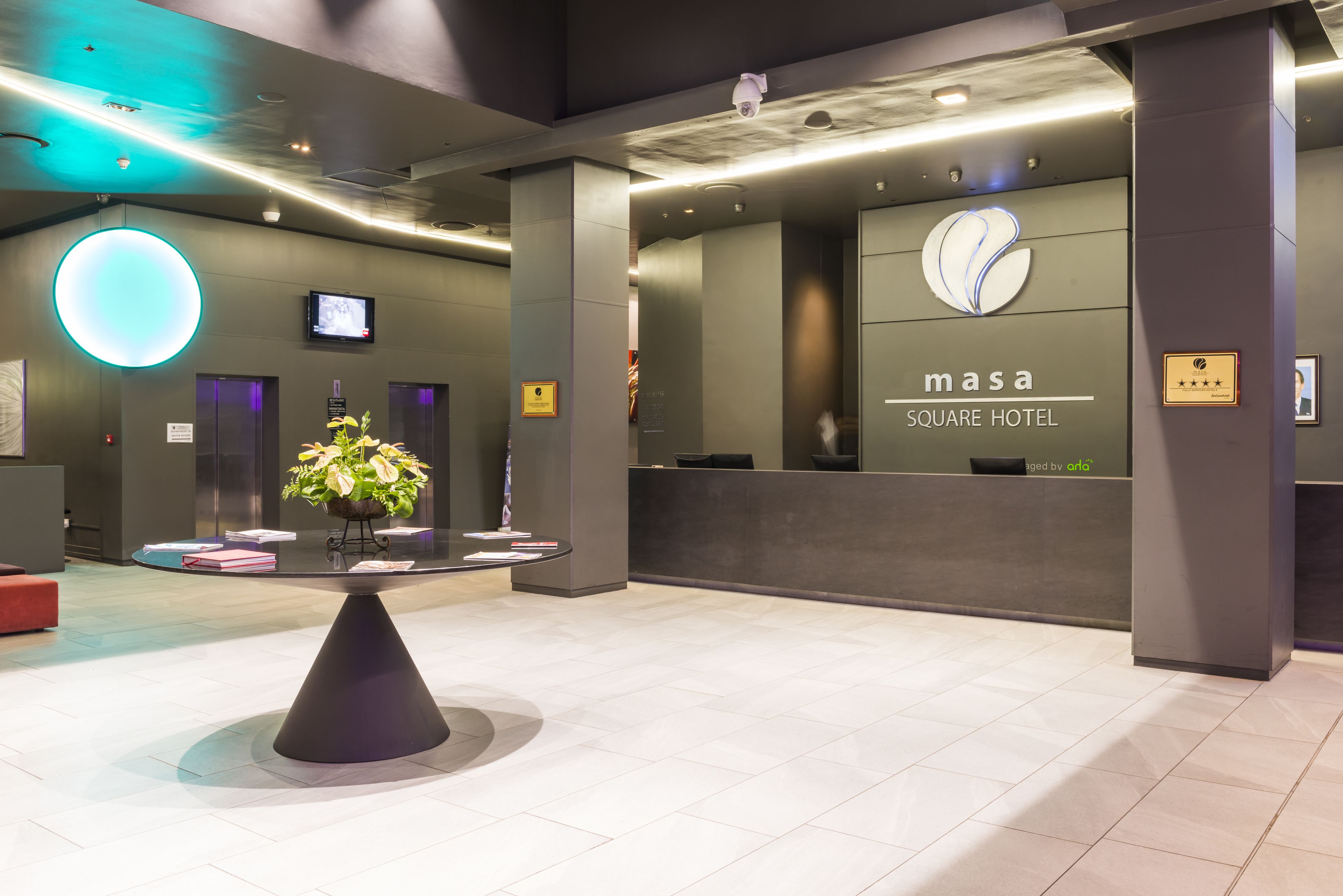 A modern hotel lobby with a reception desk, a large circular light fixture, and a potted plant.
