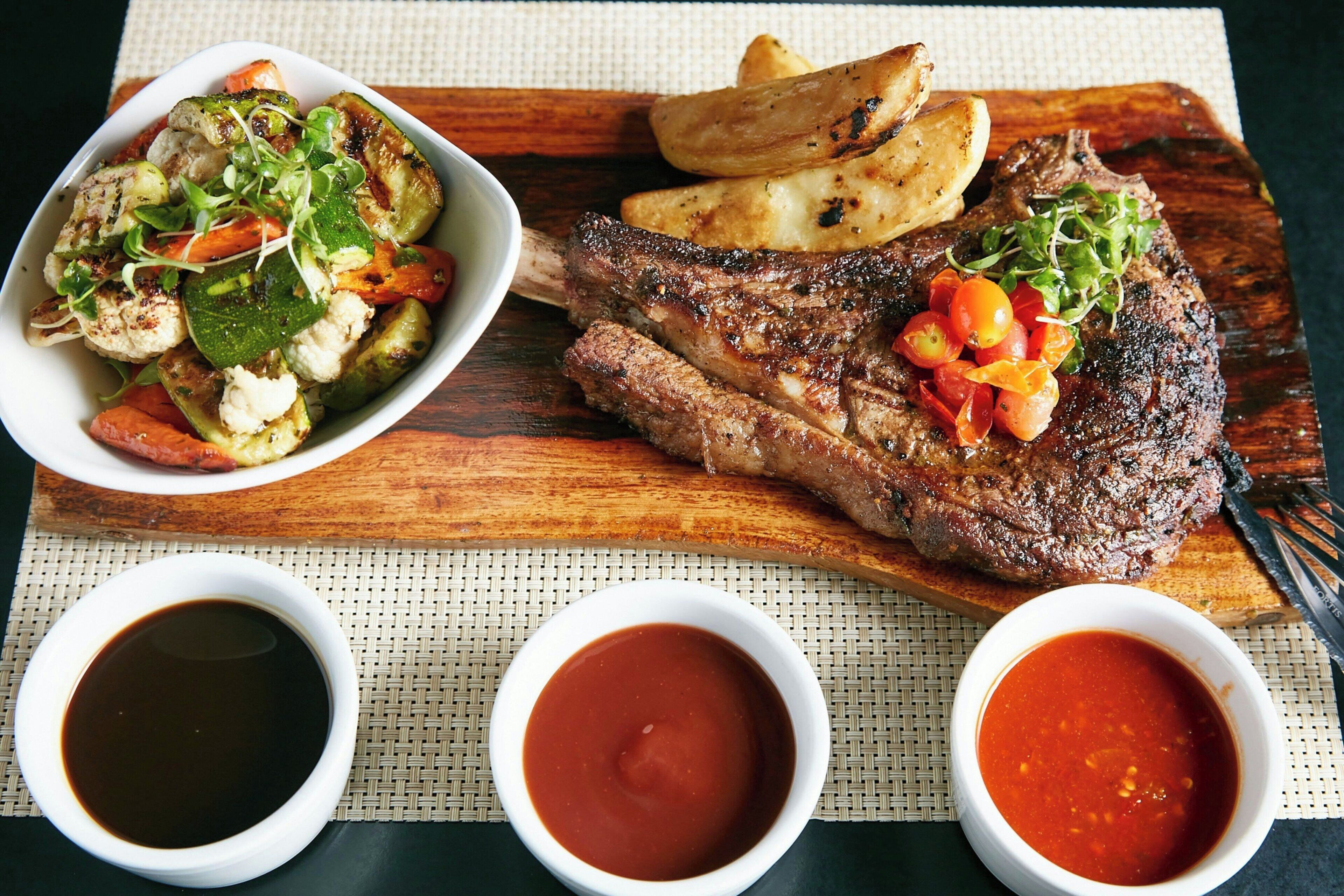 A wooden table with a steak, roasted potatoes, and a salad. There are three small bowls of sauce.