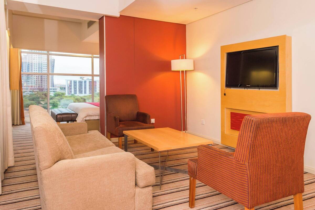 Una habitación de hotel moderna con un ventanal grande, un televisor de pantalla plana montado en la pared, un sofá, una silla y una mesita.
