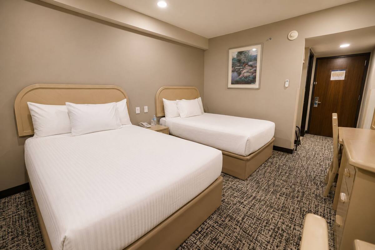 A hotel room with two beds, a desk, a chair, a painting on the wall, and a door.