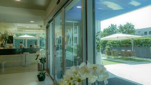 A modern hotel lobby with a reception desk, a glass door leading to an outdoor patio with umbrellas and chairs. - Kristal (Zane)