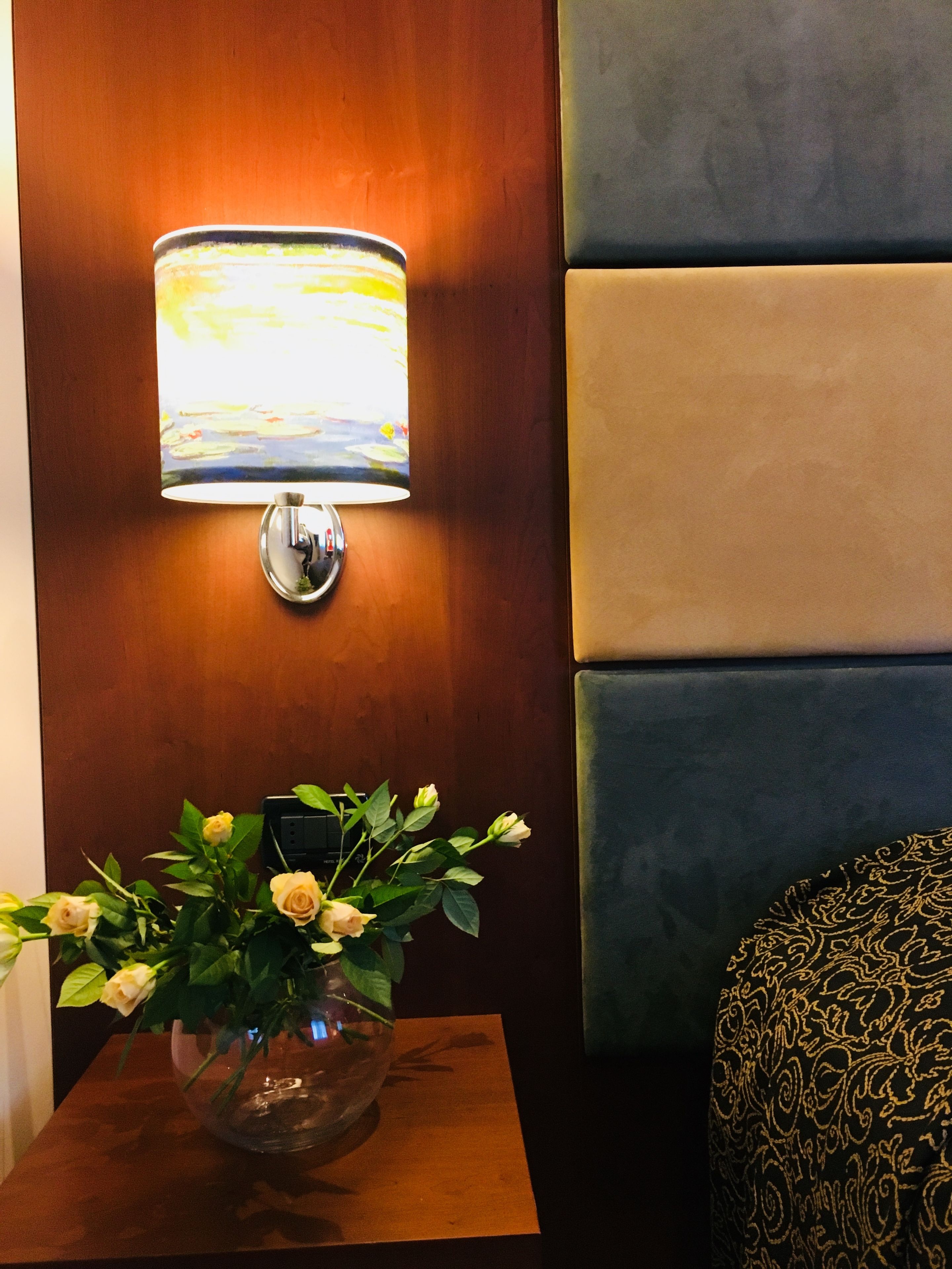 Ein Zimmer mit Wandlampe, Blumen in einer Vase und einem Bett mit gemusterten Bettwaren.