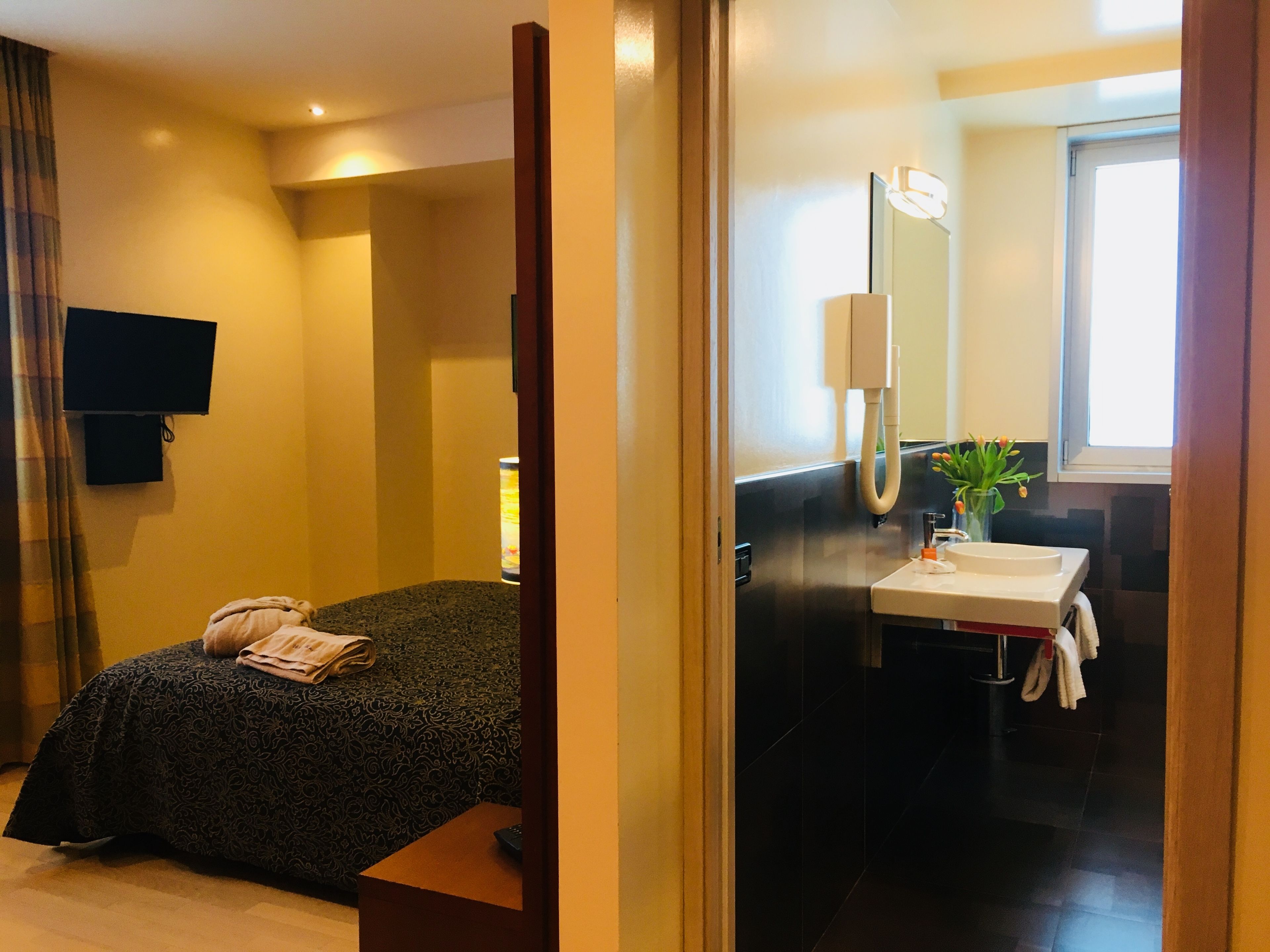 Hotelkamer met een bed, een aan de muur bevestigde televisie, een badkamer met een wastafel en spiegel, en een raam dat natuurlijk licht binnenlaat.