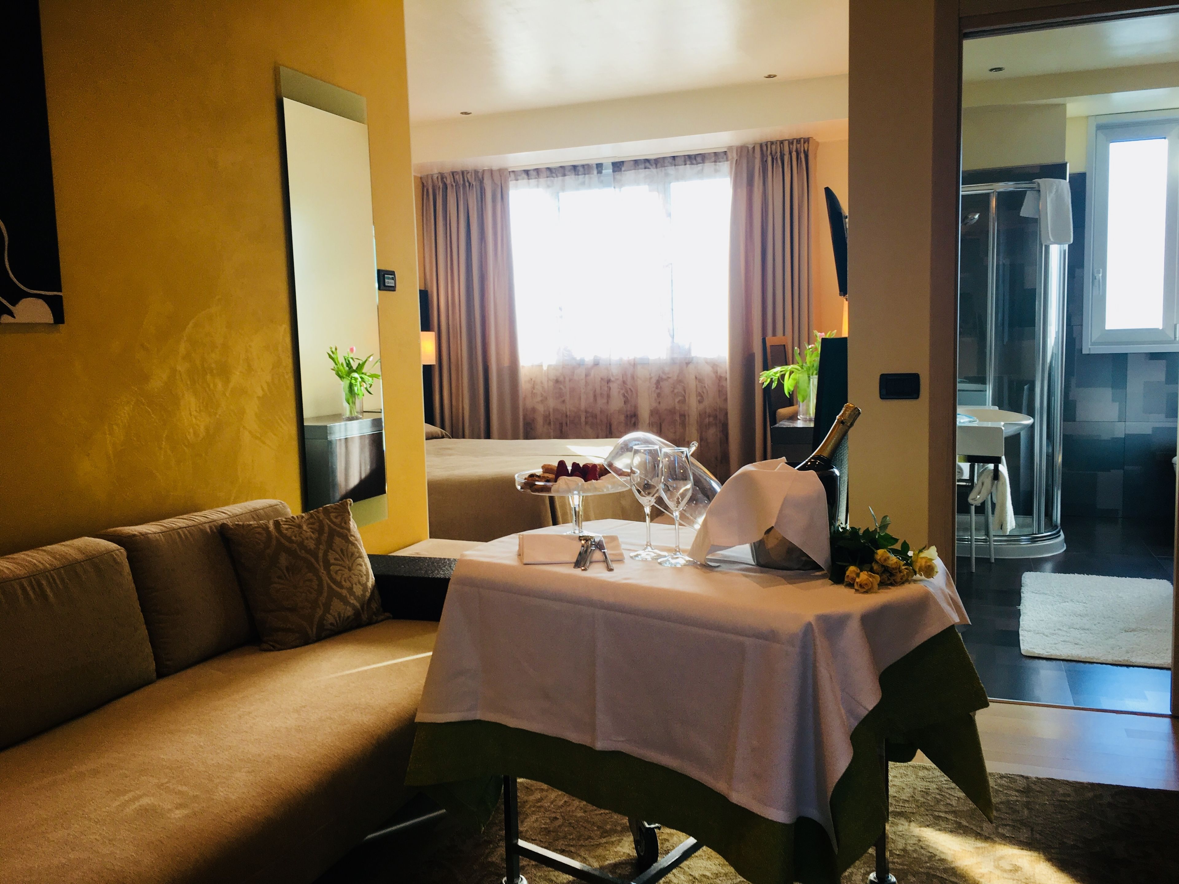 A hotel room with a sofa, a table set with a bottle of champagne, glasses, and a small plant.