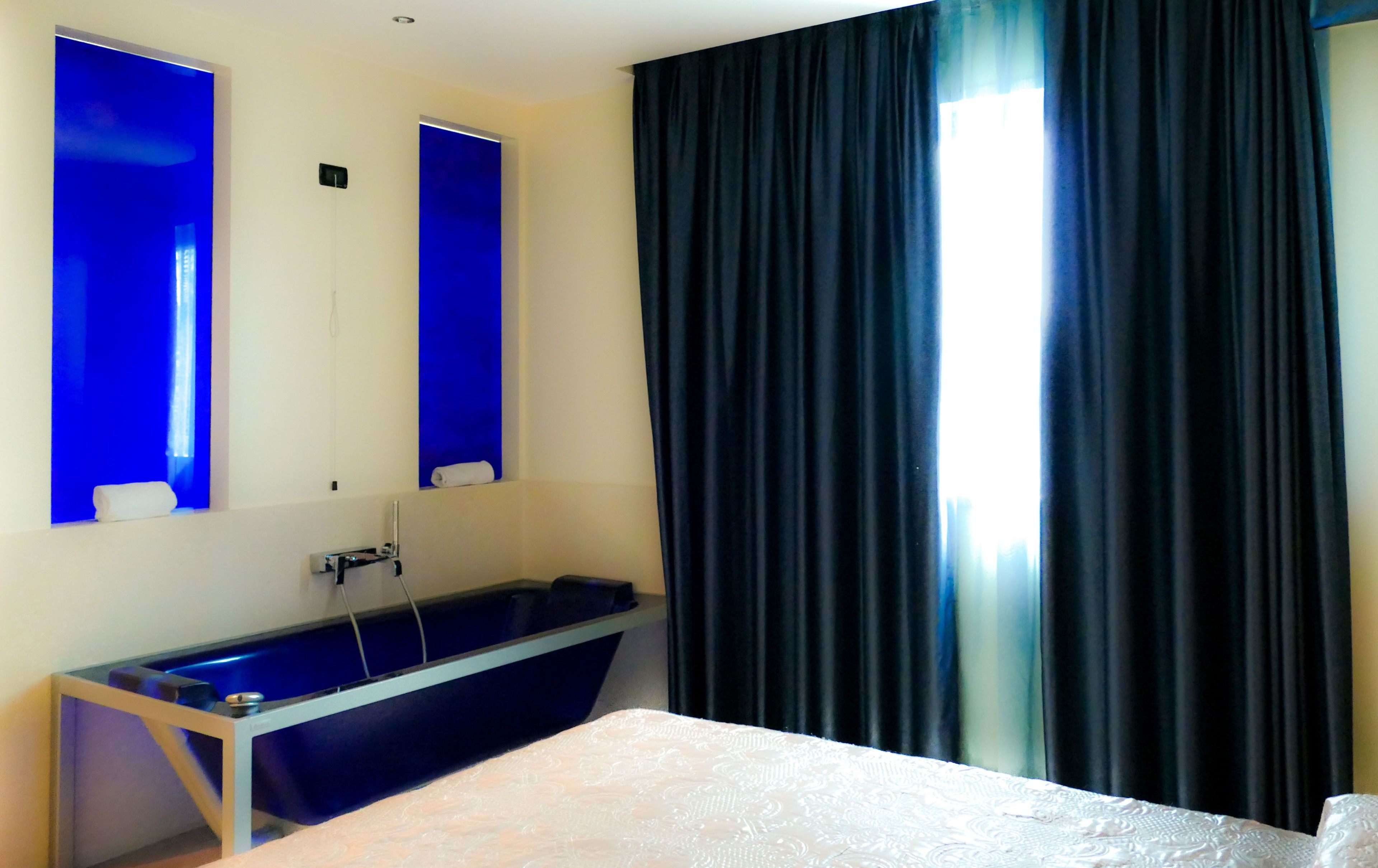 Habitación de hotel con cama, una mesita de noche, lavamanos integrado y un ventanal grande con cortinas oscuras.