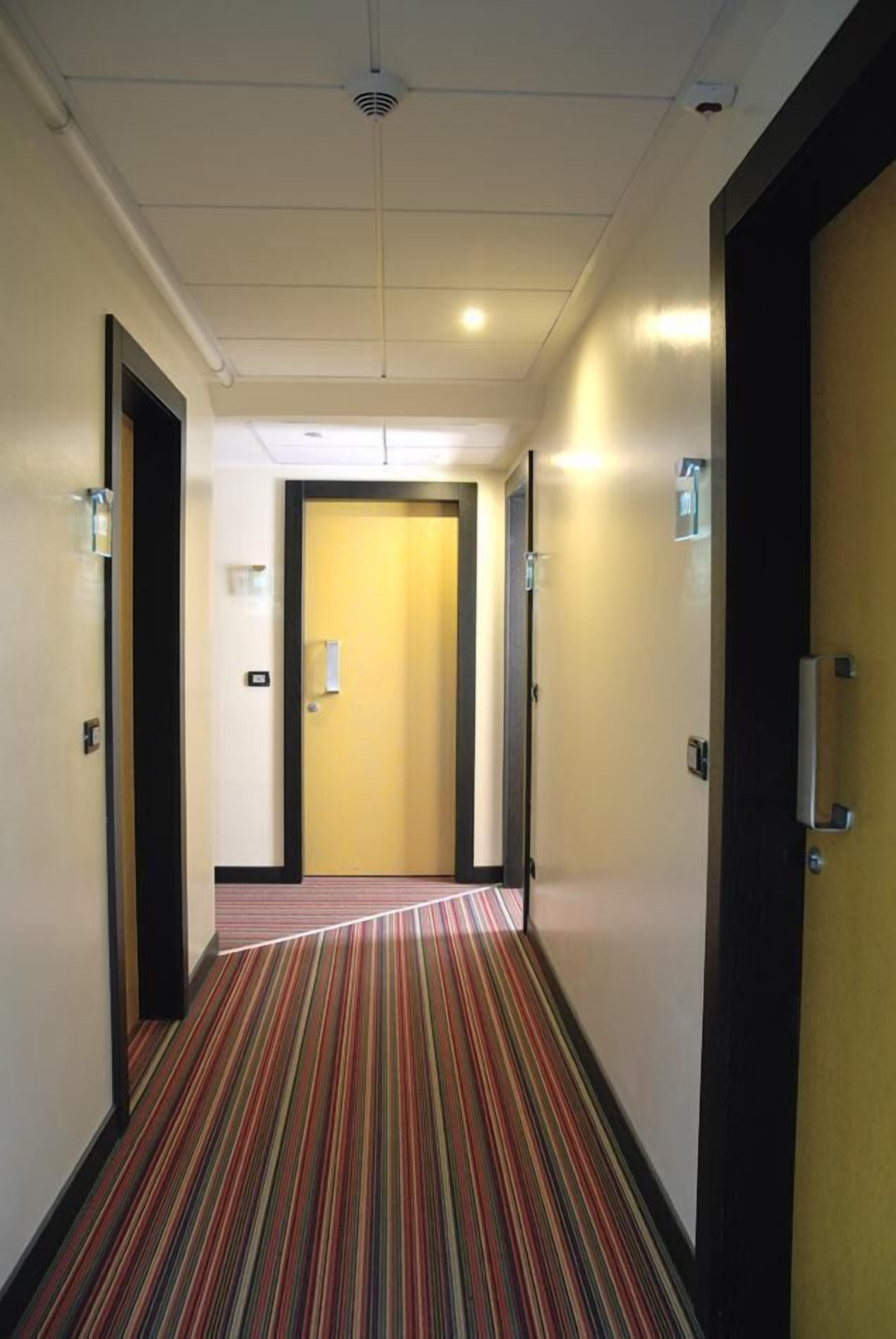 Un couloir d'hôtel avec un tapis à rayures, des portes et un luminaire au plafond.