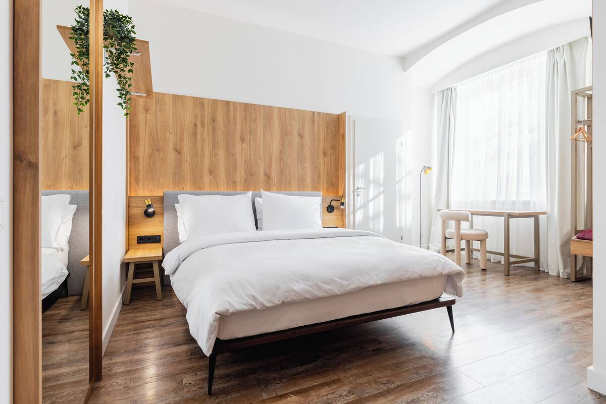 A modern hotel room with a wooden headboard, a large bed, a small desk, and a chair.