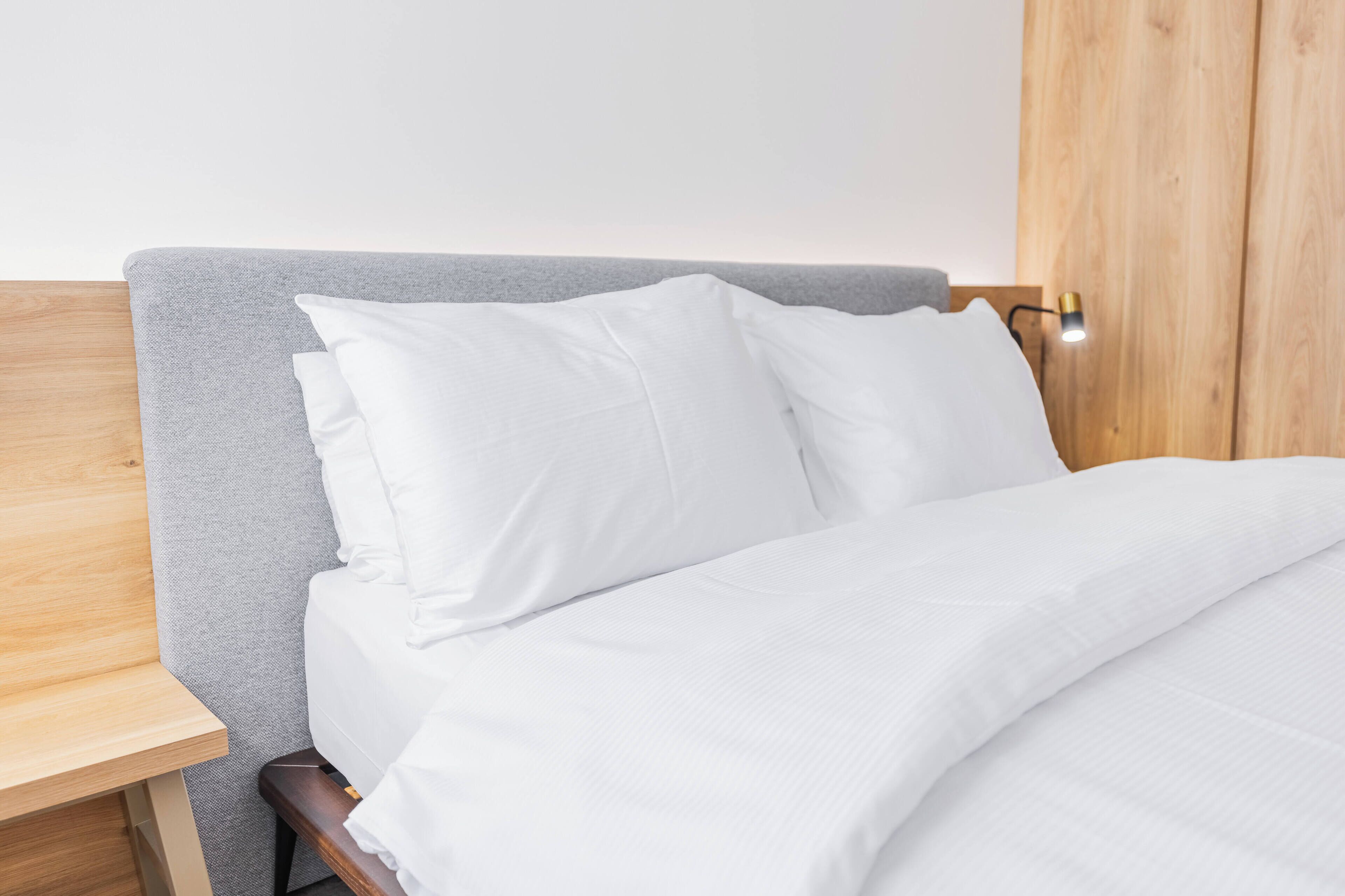 Un lit bien fait, avec du linge de lit blanc et des oreillers, une table de chevet en bois et un luminaire fixé au mur.