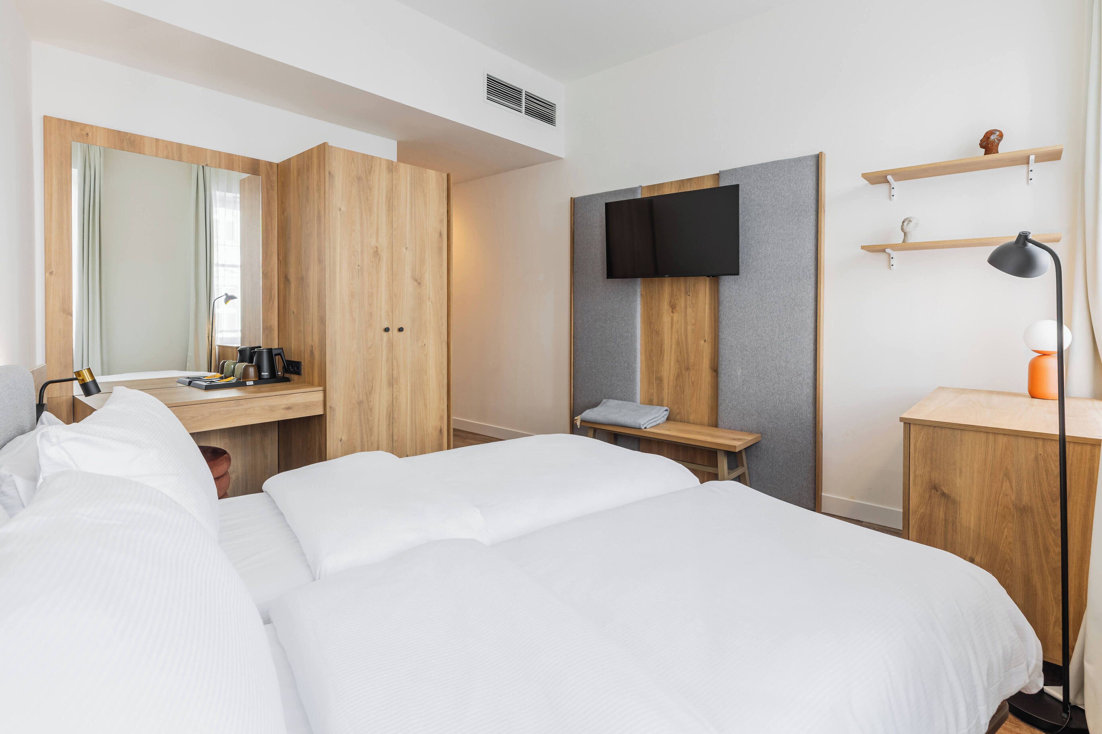 A hotel room with a large bed, a wooden wardrobe, a desk with a flat-screen TV, and a small table with a lamp.