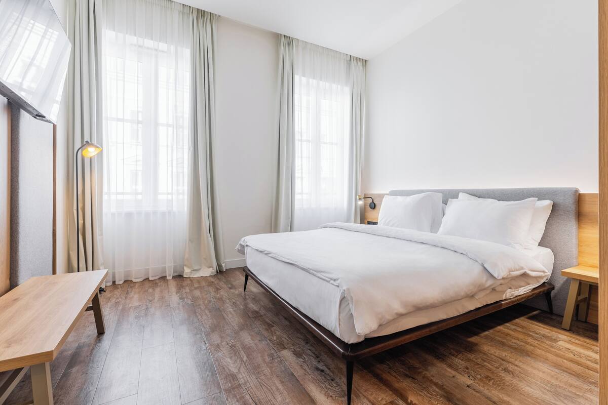 A bedroom with a wooden floor, a bed with white bedding, a bedside table, a lamp, and a window with sheer curtains.