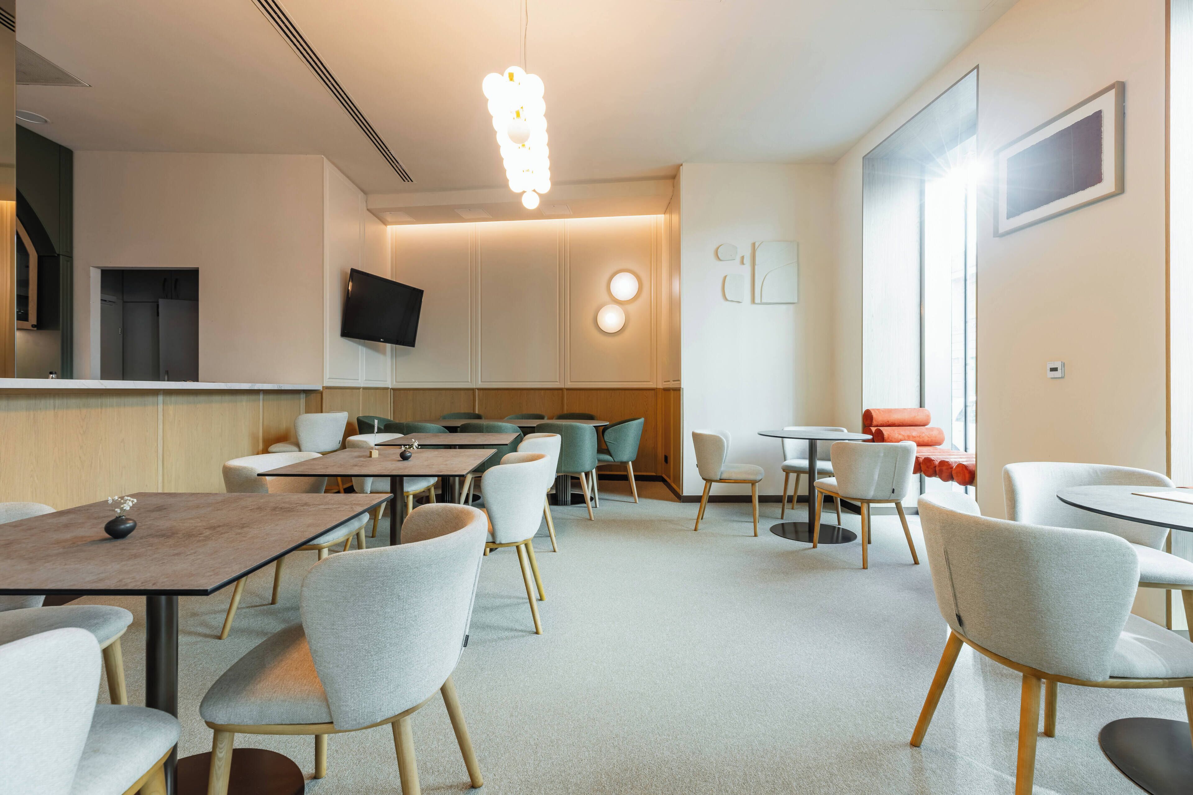A modern dining area with tables and chairs, a flat-screen TV mounted on the wall, and a large window providing natural light.