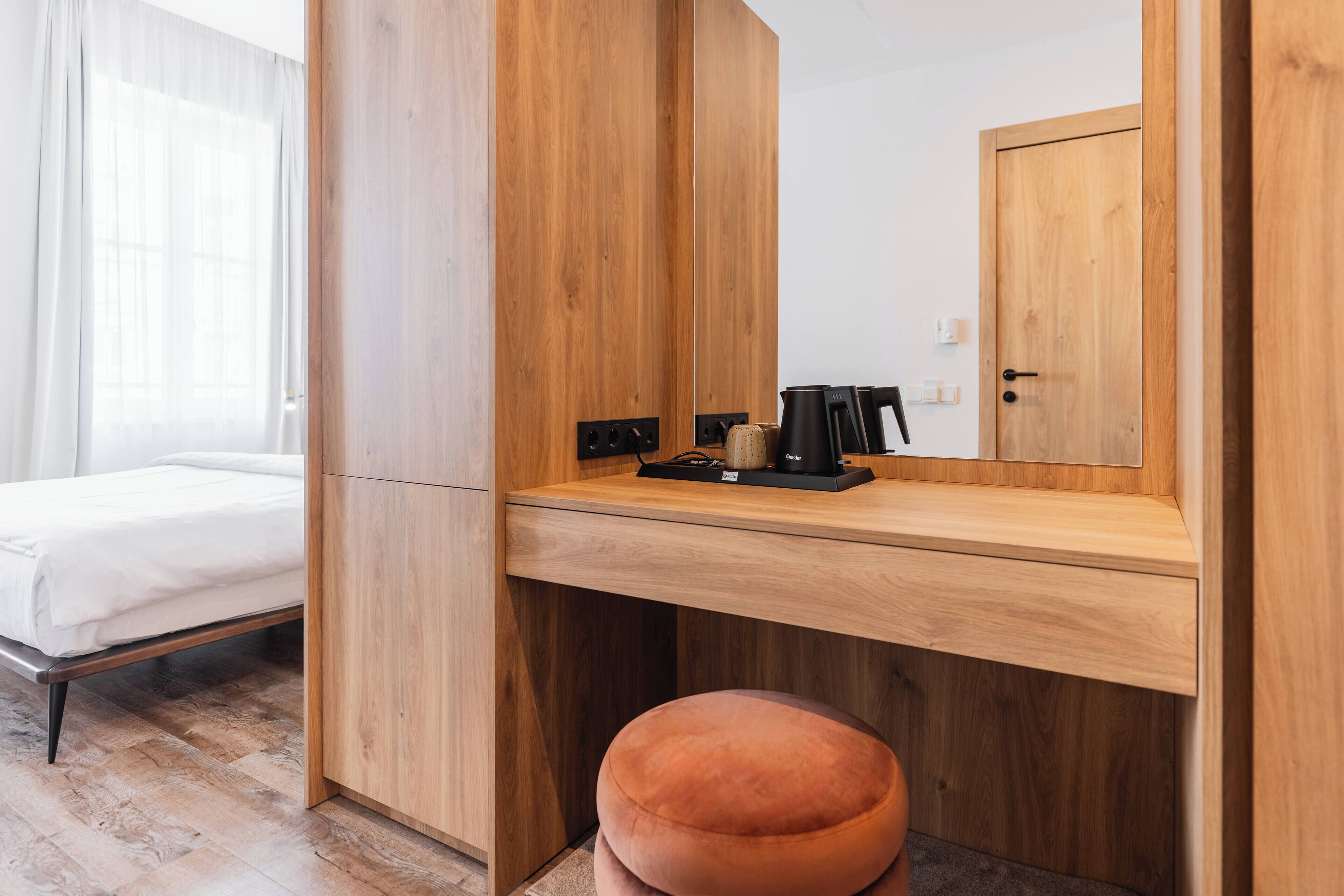 A modern room with a wooden desk, a mirror, a bed, and a small orange ottoman.